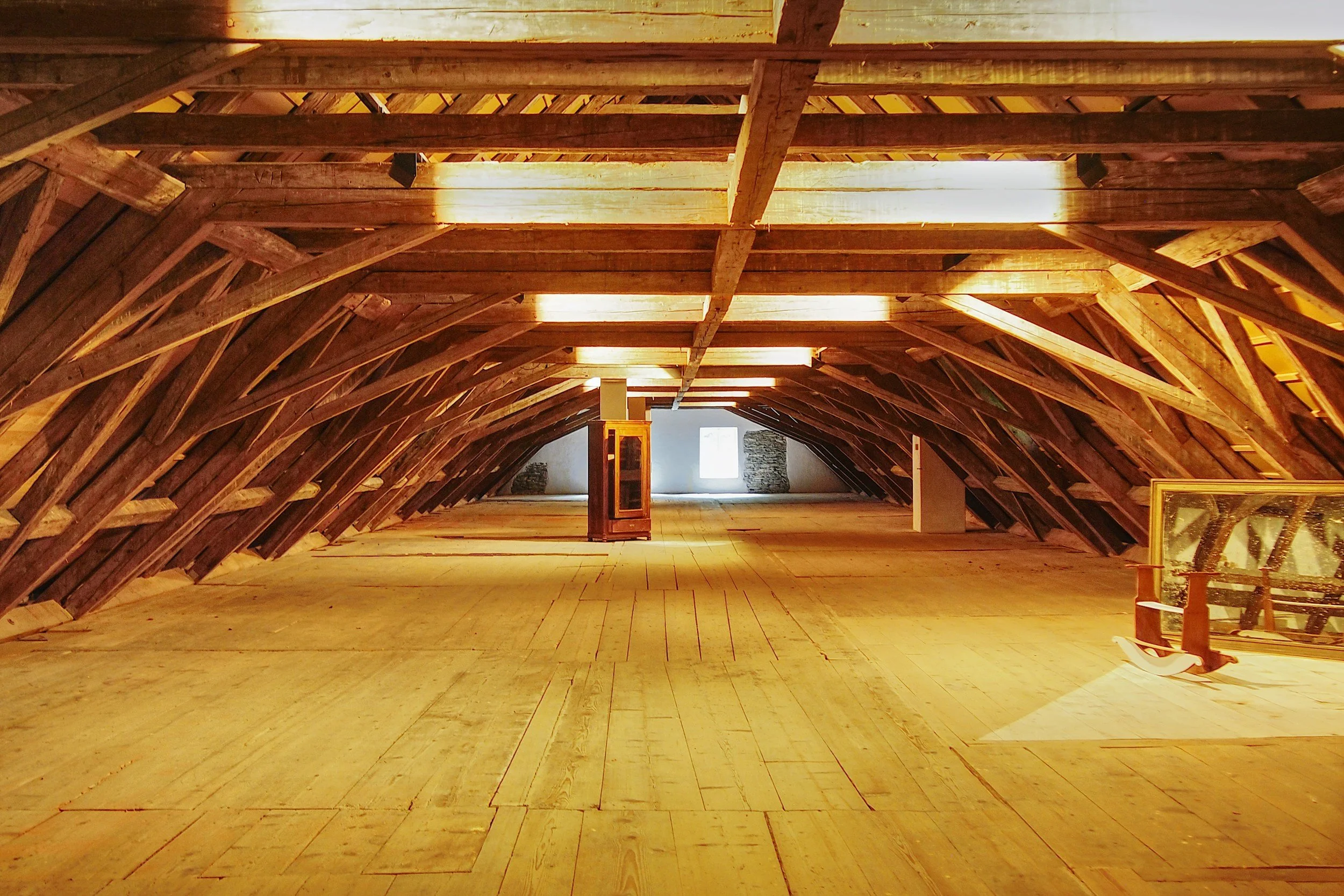Interior view of an attic with exposed wooden beams, sloped ceiling, and a natural light source at the far end.
