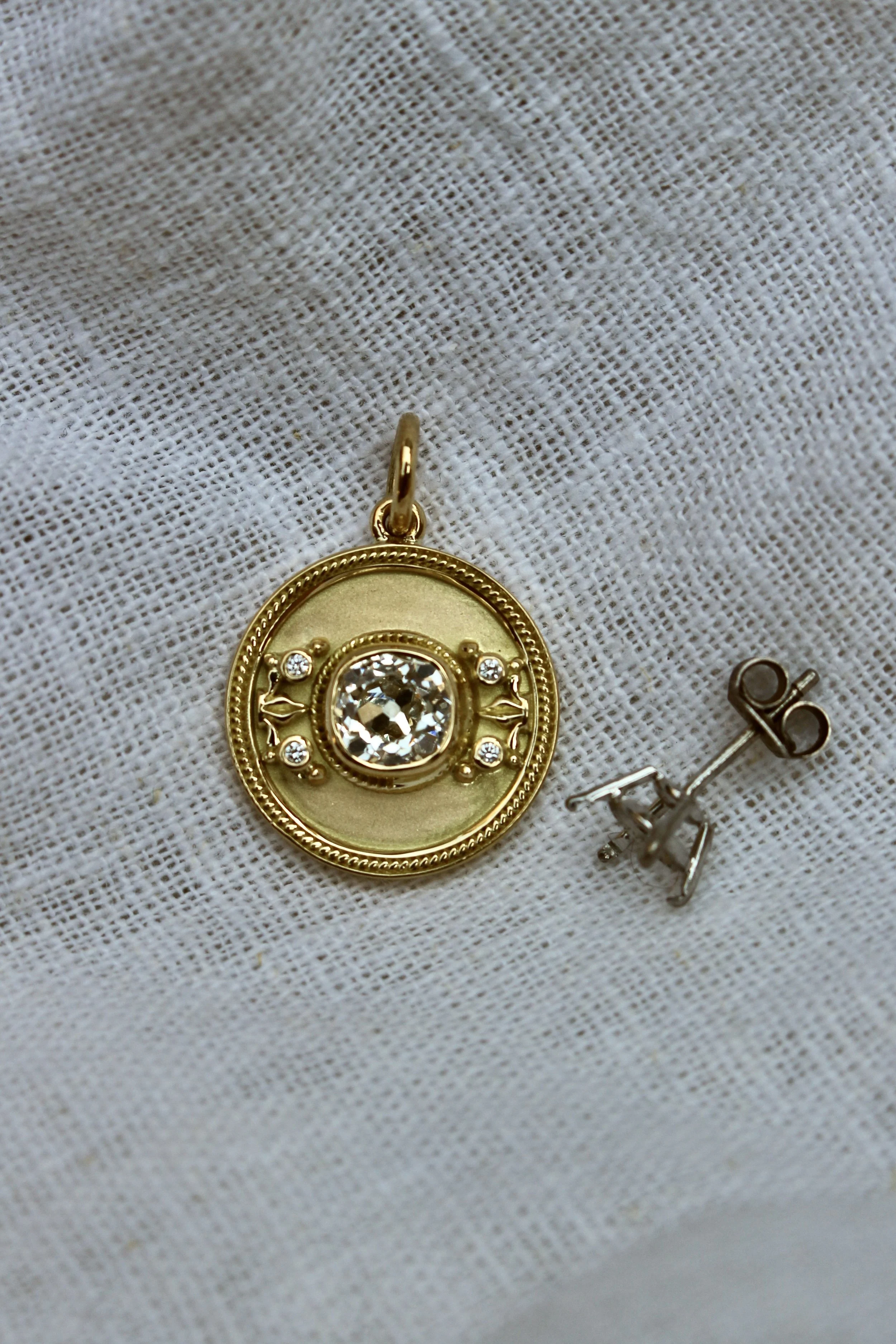 Golden locket with a large central diamond and smaller diamonds, resting on white textured fabric, with a pair of small silver earrings nearby.