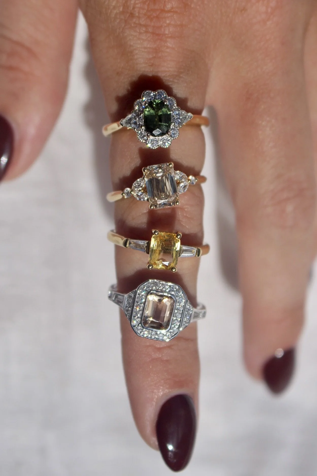 Close-up of a hand with multiple rings featuring various gemstones and diamonds, with dark red nail polish.