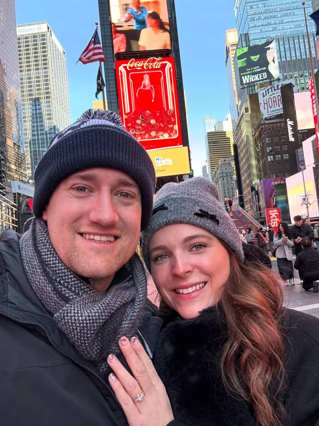 A couple taking a selfie in Times Square, New York City, wearing winter hats and scarves, with bright billboards and buildings in the background.