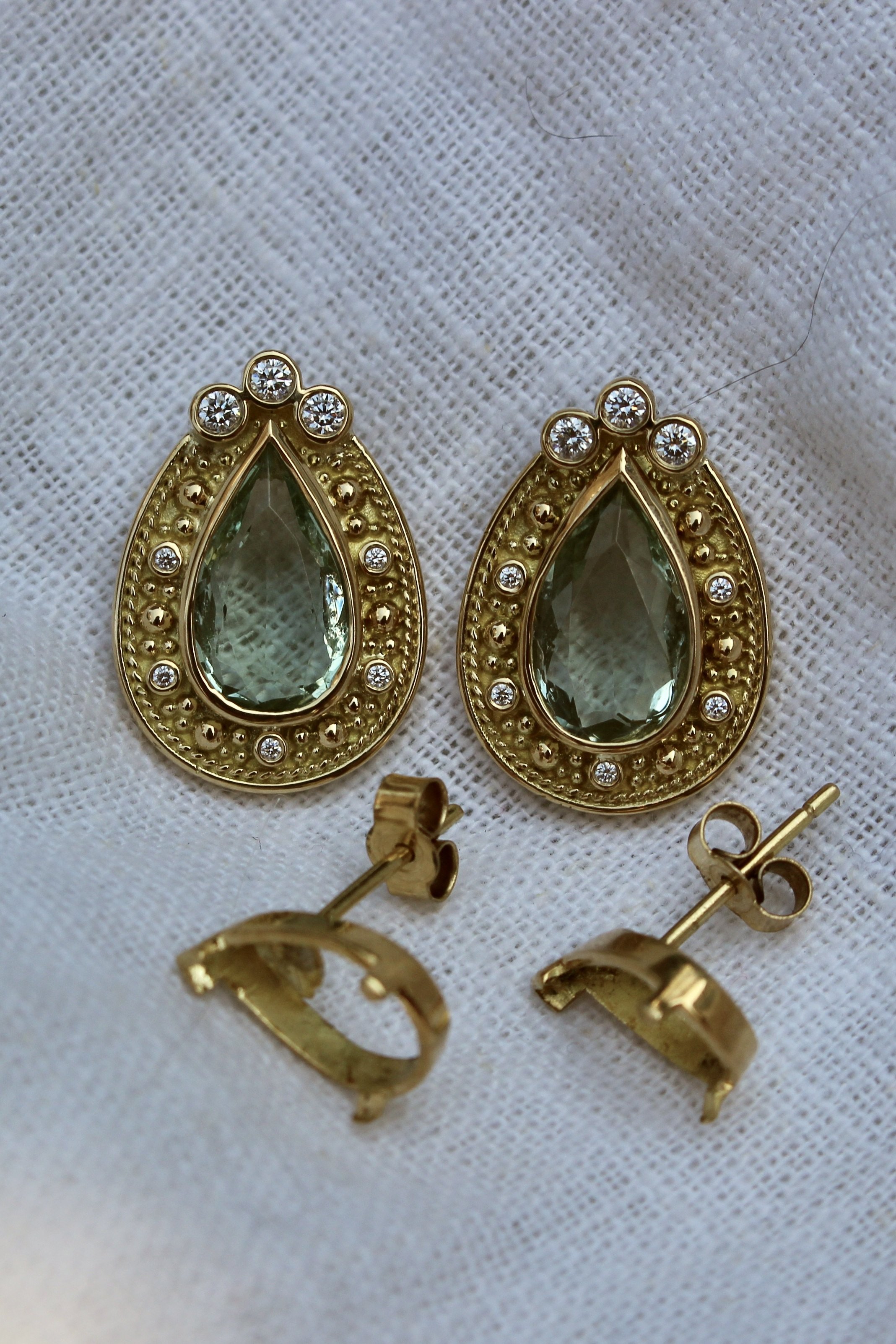Gold earrings with teardrop-shaped green gemstones and small white rhinestones, placed on a white textured fabric background.