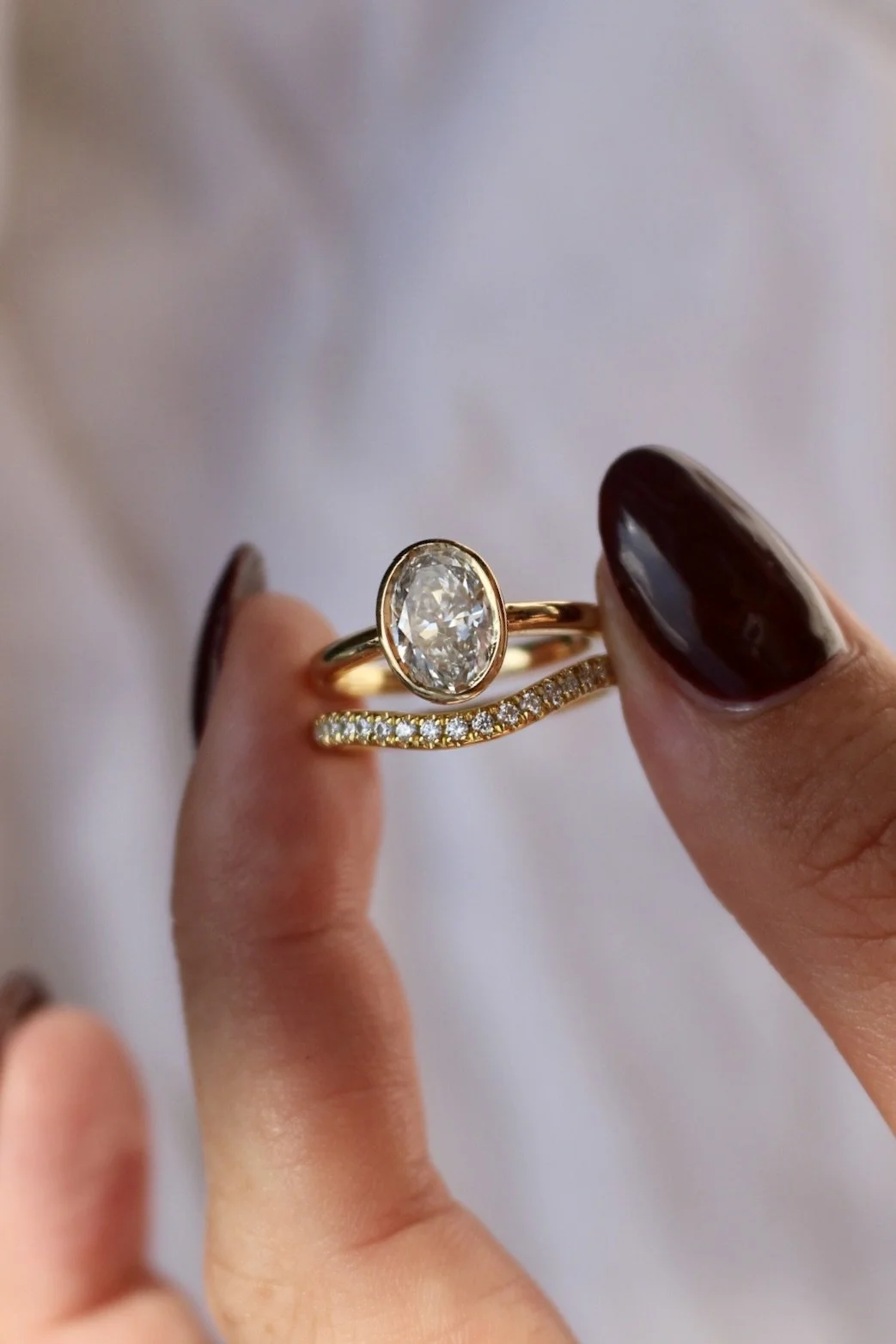 Close-up of two gold rings, one with an oval diamond and the other with small diamonds along the band, held between a person's fingers with dark nail polish.