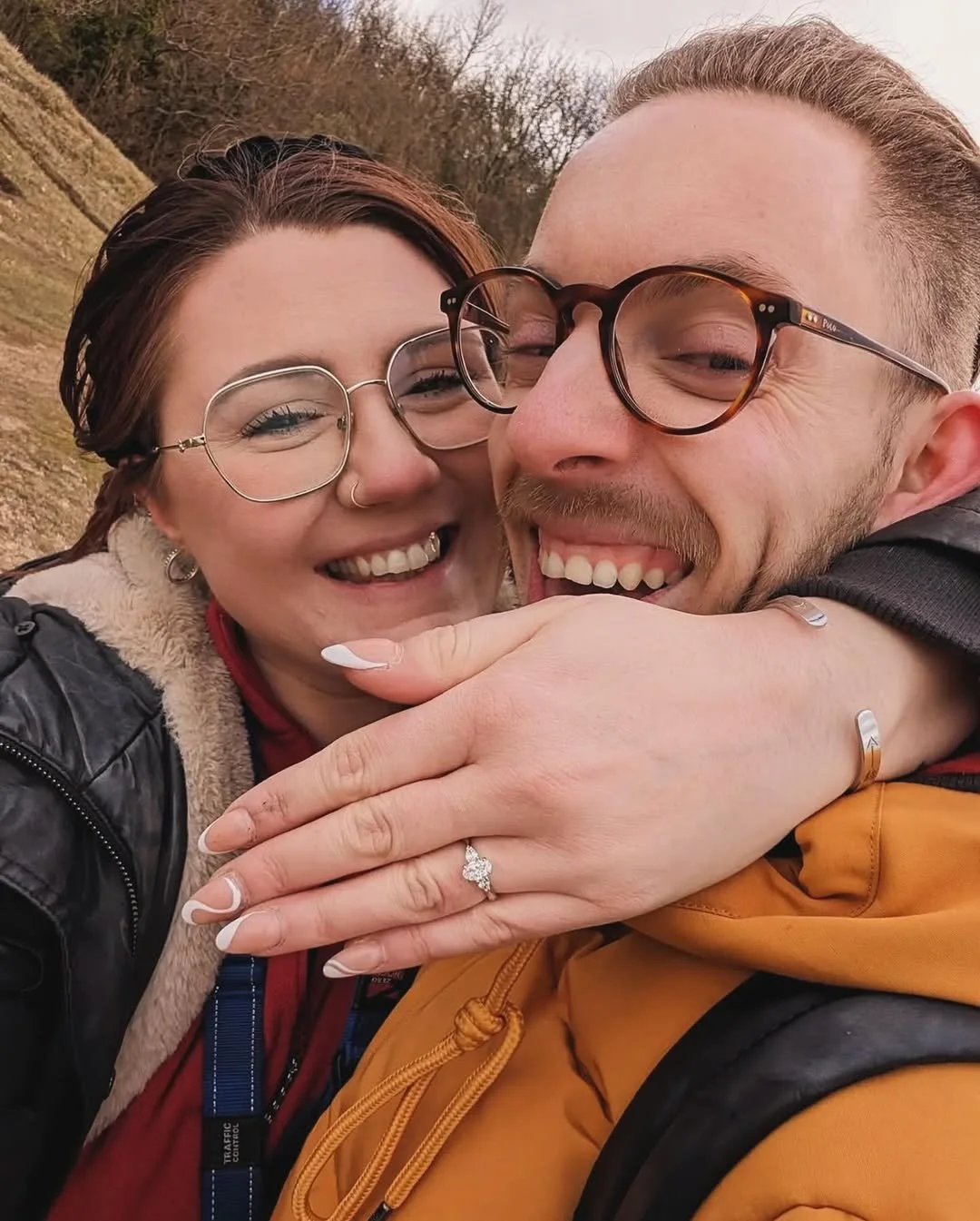 A smiling couple outdoors, hugging each other tightly. The woman has red hair, glasses, and a nose ring, and is wearing a black jacket with a fur collar. The man has light brown hair, glasses, and a beard, and is wearing a yellow hoodie. The woman's hand with a ring is visible on the man's chest, and they are surrounded by a natural background with trees and grass.