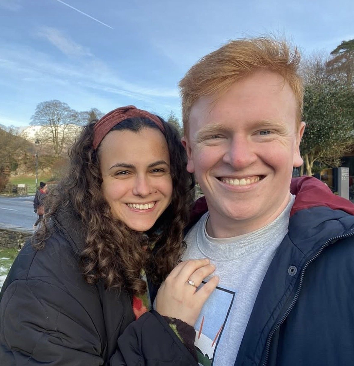 A smiling couple taking a selfie outdoors during daytime with trees, a stone wall, and a clear blue sky in the background.