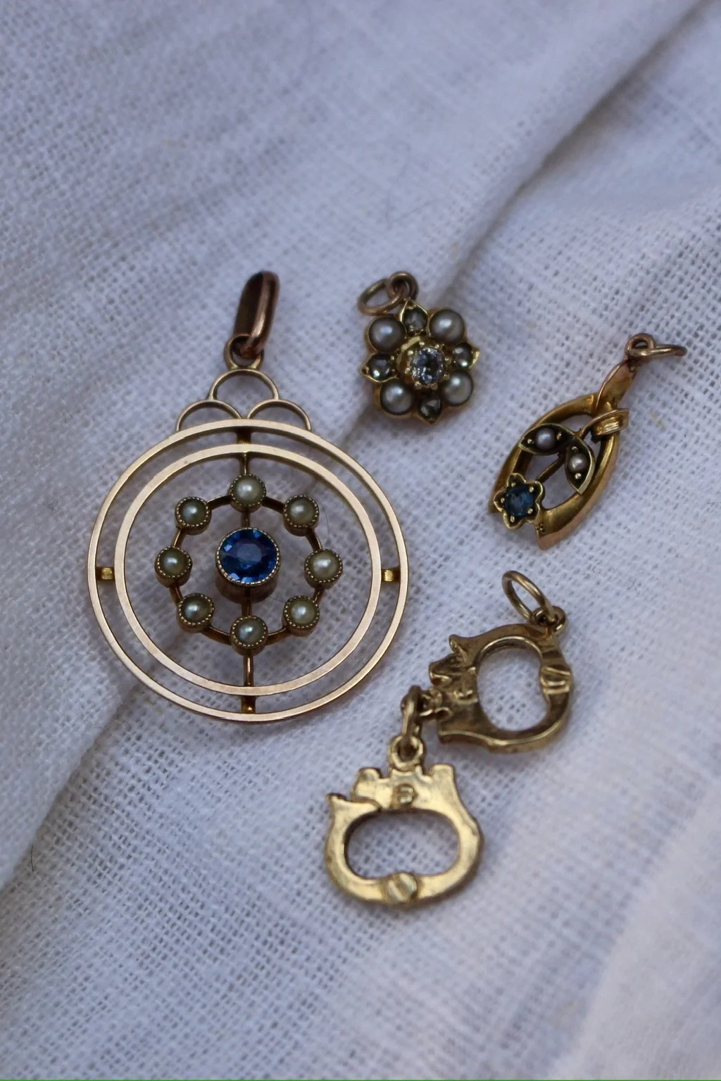 Four gold and pearl jewelry charms on a white fabric background, including a large circular pendant with pearls and a blue gemstone, a small floral charm with pearls and a clear gemstone, a teardrop-shaped charm with pearls and a small blue gemstone,