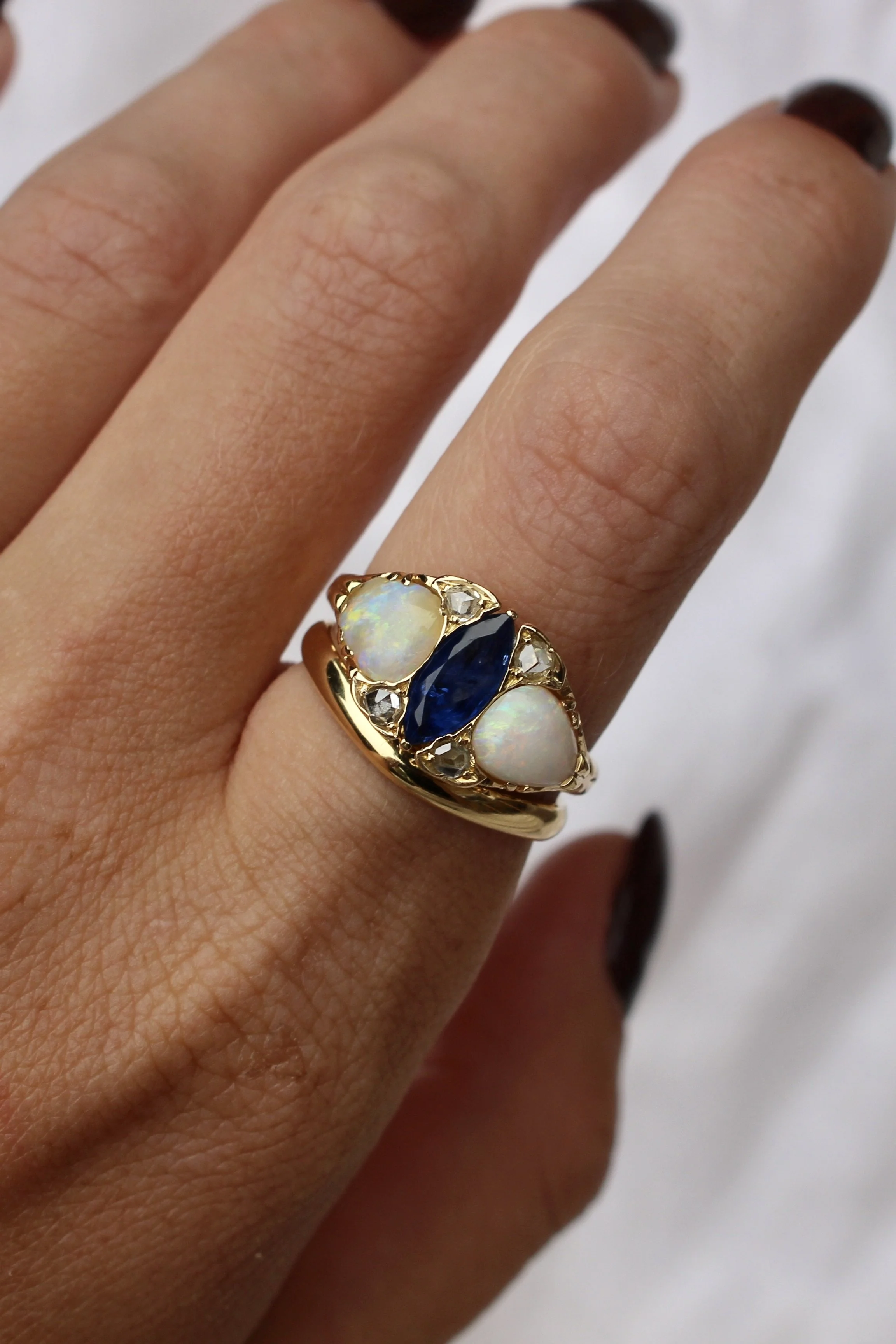 Close-up of a hand wearing a gold ring with opal and blue gemstone, with dark painted nails.