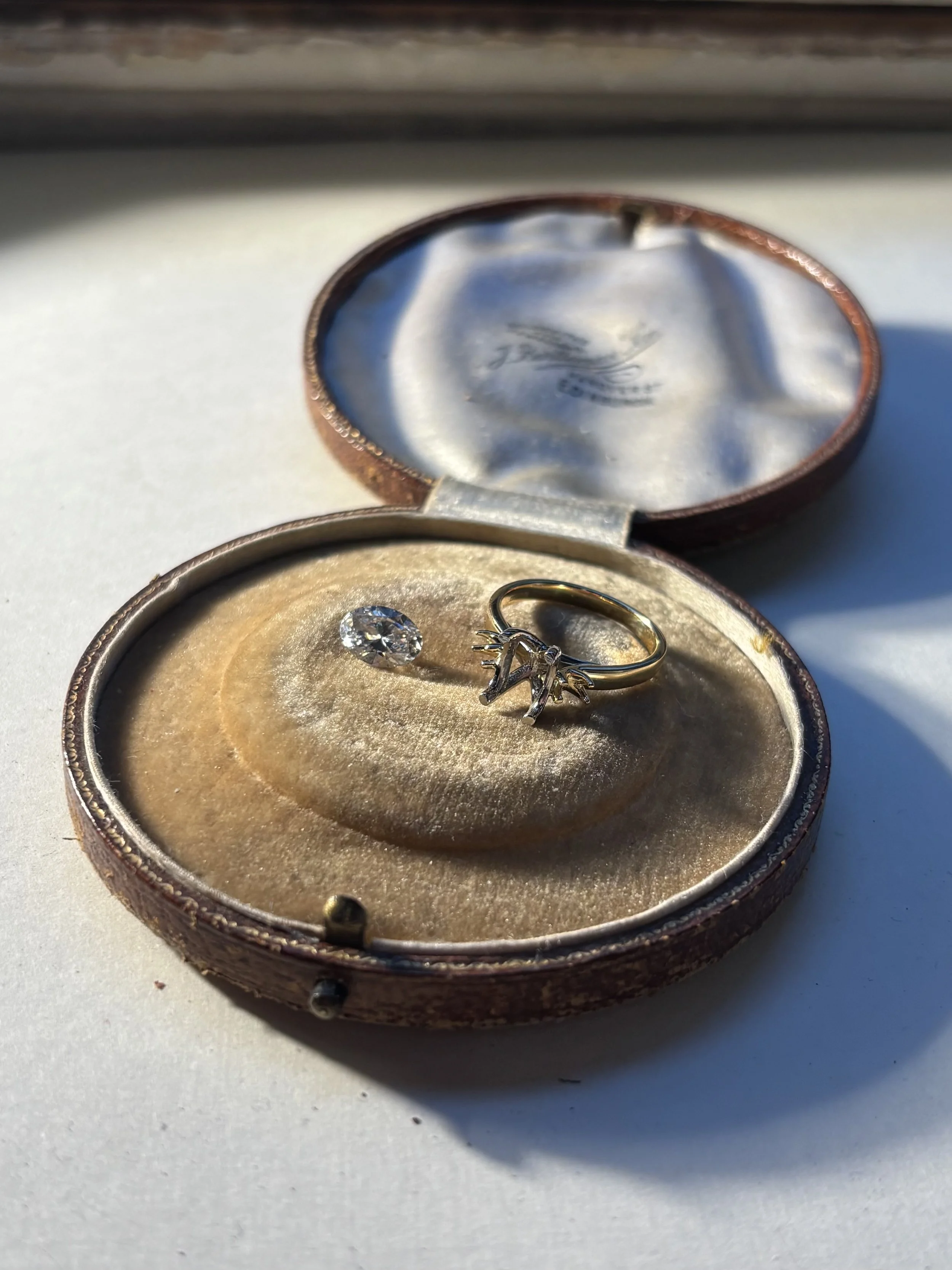 A small oval jewelry box with a ring and a diamond inside, placed on a white surface. The box has a velvet interior and a metallic exterior.