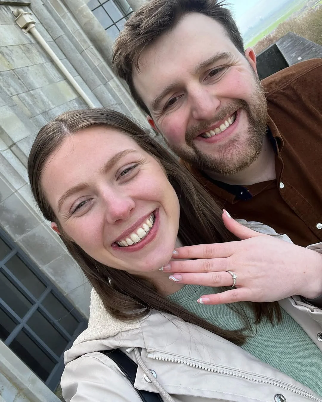 A smiling young woman with long brown hair and a ring on her finger, and a smiling young man with a beard and brown shirt, taking a selfie outdoors near a stone building.