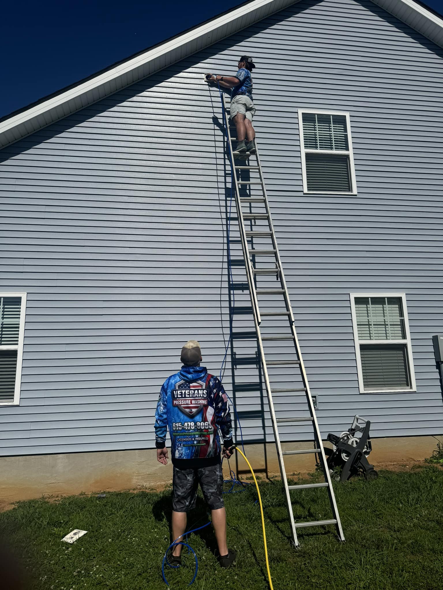 A person standing on the ground holding a hose, watching another person on a ladder cleaning siding of a light blue house.