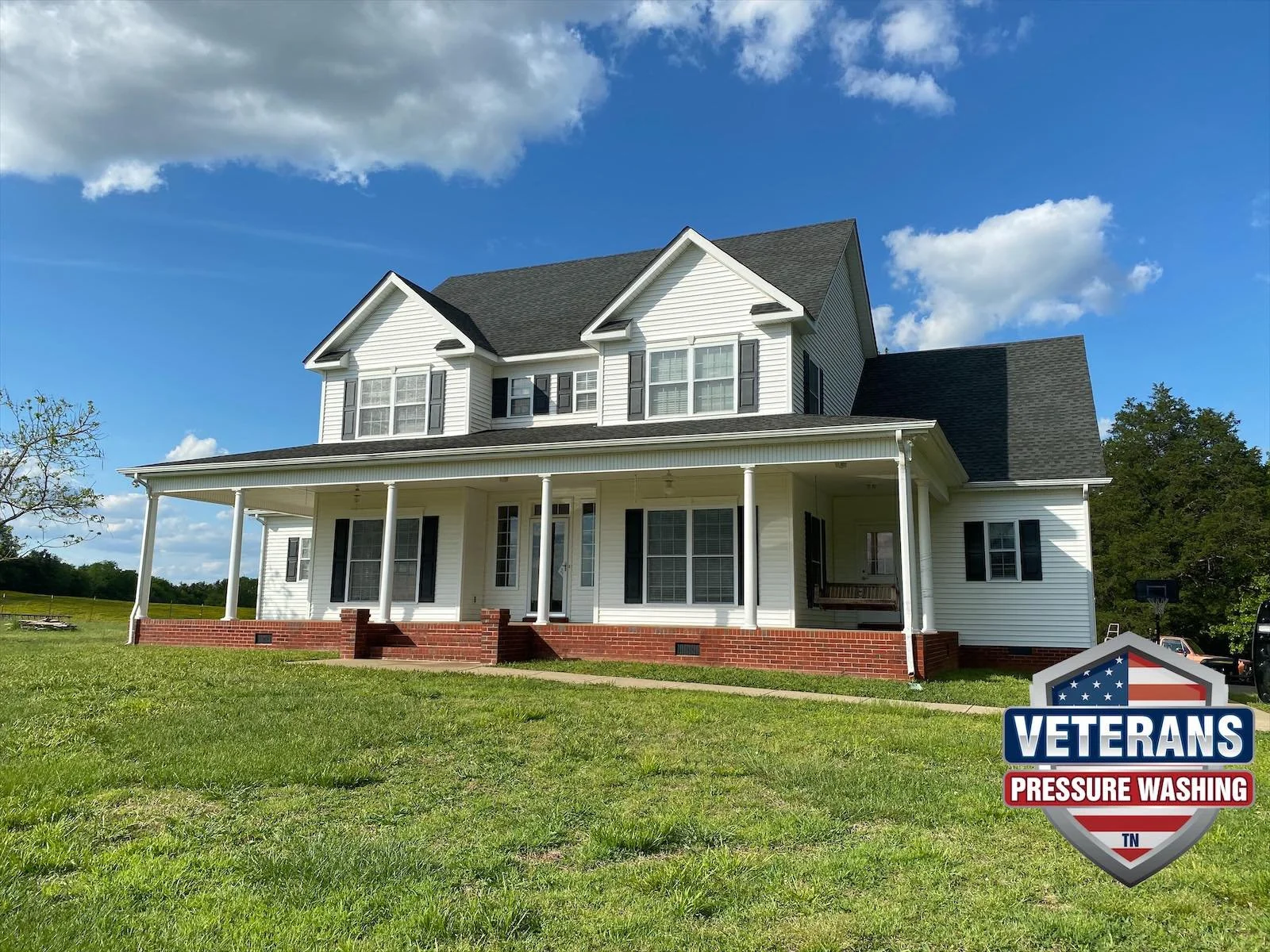 A large white two-story house with black shutters and a covered front porch with white columns, surrounded by green grass and a clear blue sky with a few clouds. There is a veterns pressure washing logo at the bottom right corner.