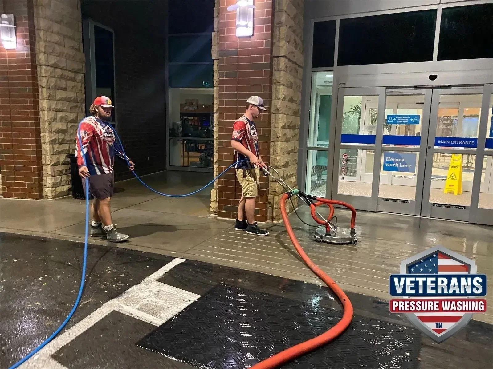 Two veterans wearing patriotic shirts and shorts pressure washing the entrance of a building at night. One veteran is operating a pressure washer, while the other holds the hose. The scene is outside a commercial building with glass doors and a sign that reads "Heroes work here". There is a black doormat and wet pavement.