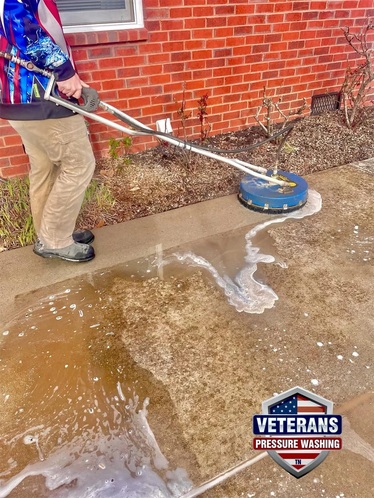 Person using a pressure washer to clean a concrete sidewalk next to a brick wall, with a logo that says 'Veterans Pressure Washing'.