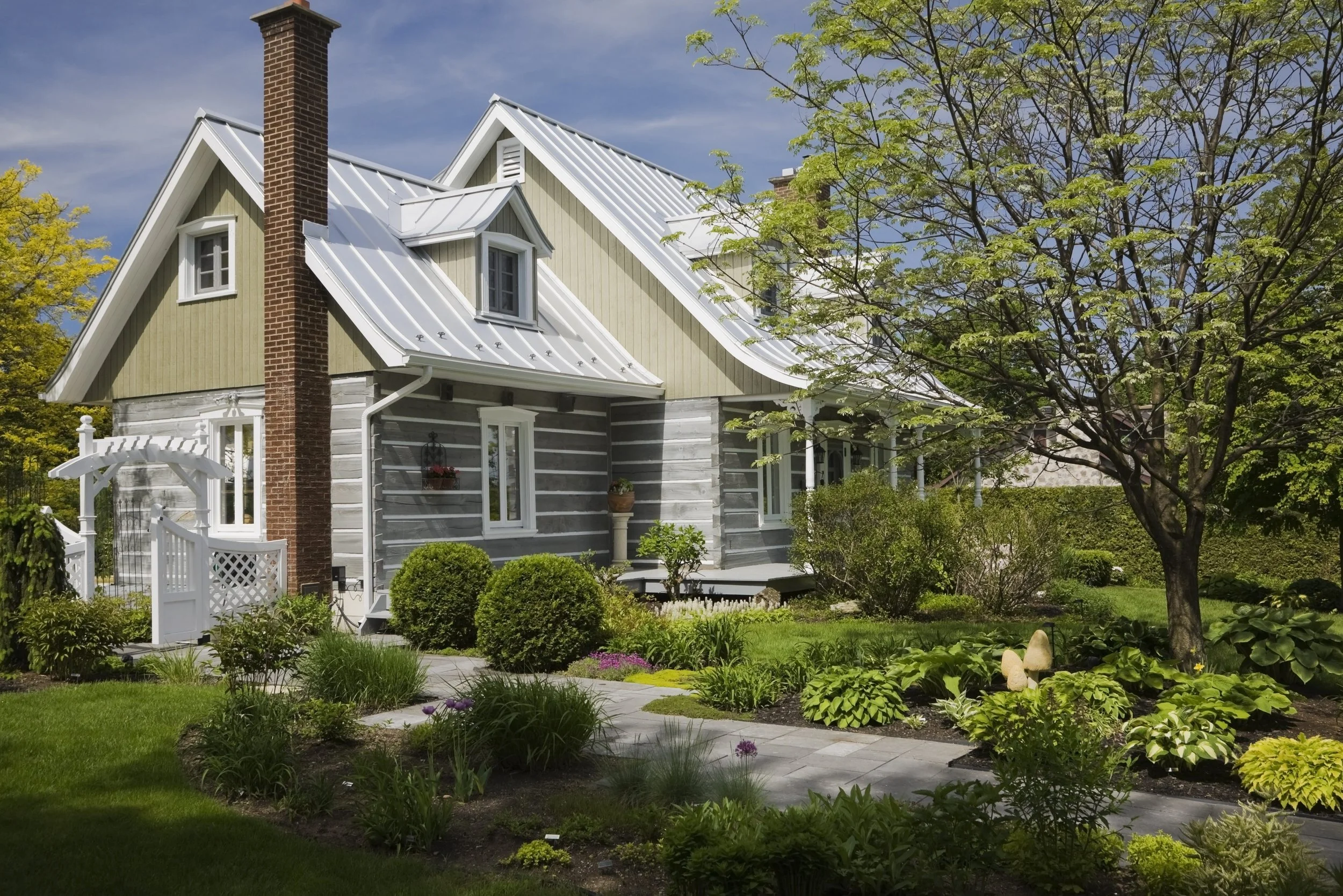 A charming house with a gray and beige exterior, white window frames, and a white metal roof. It is surrounded by a well-maintained garden with green bushes, a flowering tree, and a stone pathway.