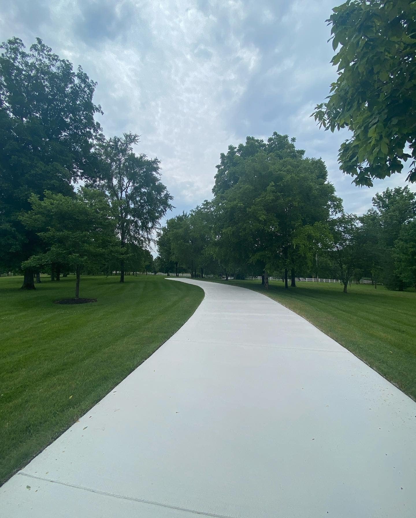 A winding concrete pathway in a park, bordered by green grass and trees, with a cloudy sky overhead.
