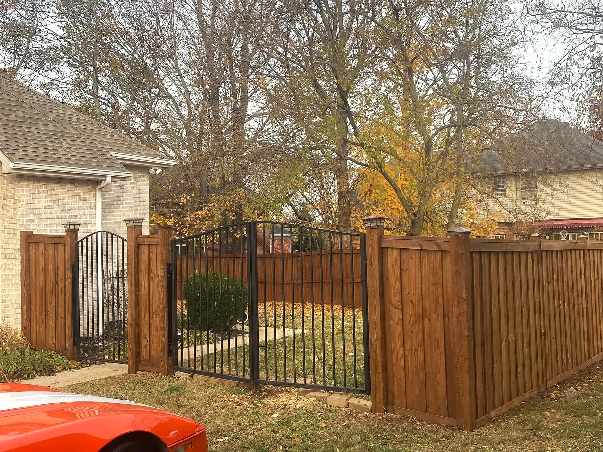 A backyard view showing a wooden fence with black iron gate, trees with autumn leaves, and neighboring houses with brick and siding exteriors.