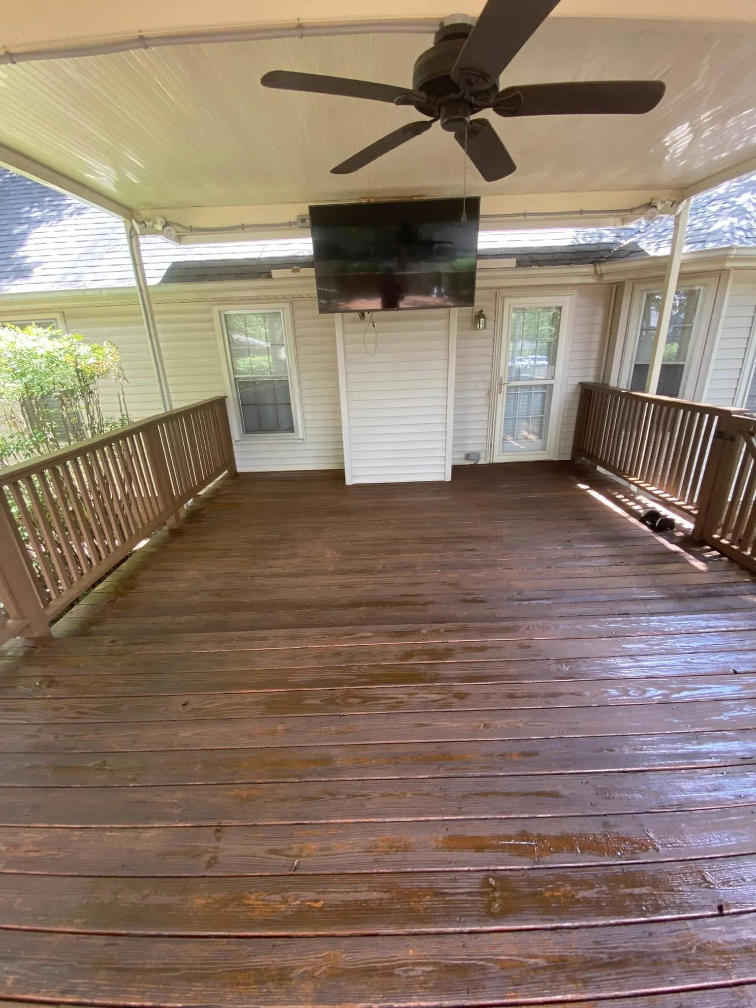 Covered outdoor porch with wooden flooring, railing, ceiling fan, and mounted TV, with house exterior, windows, and door in the background.