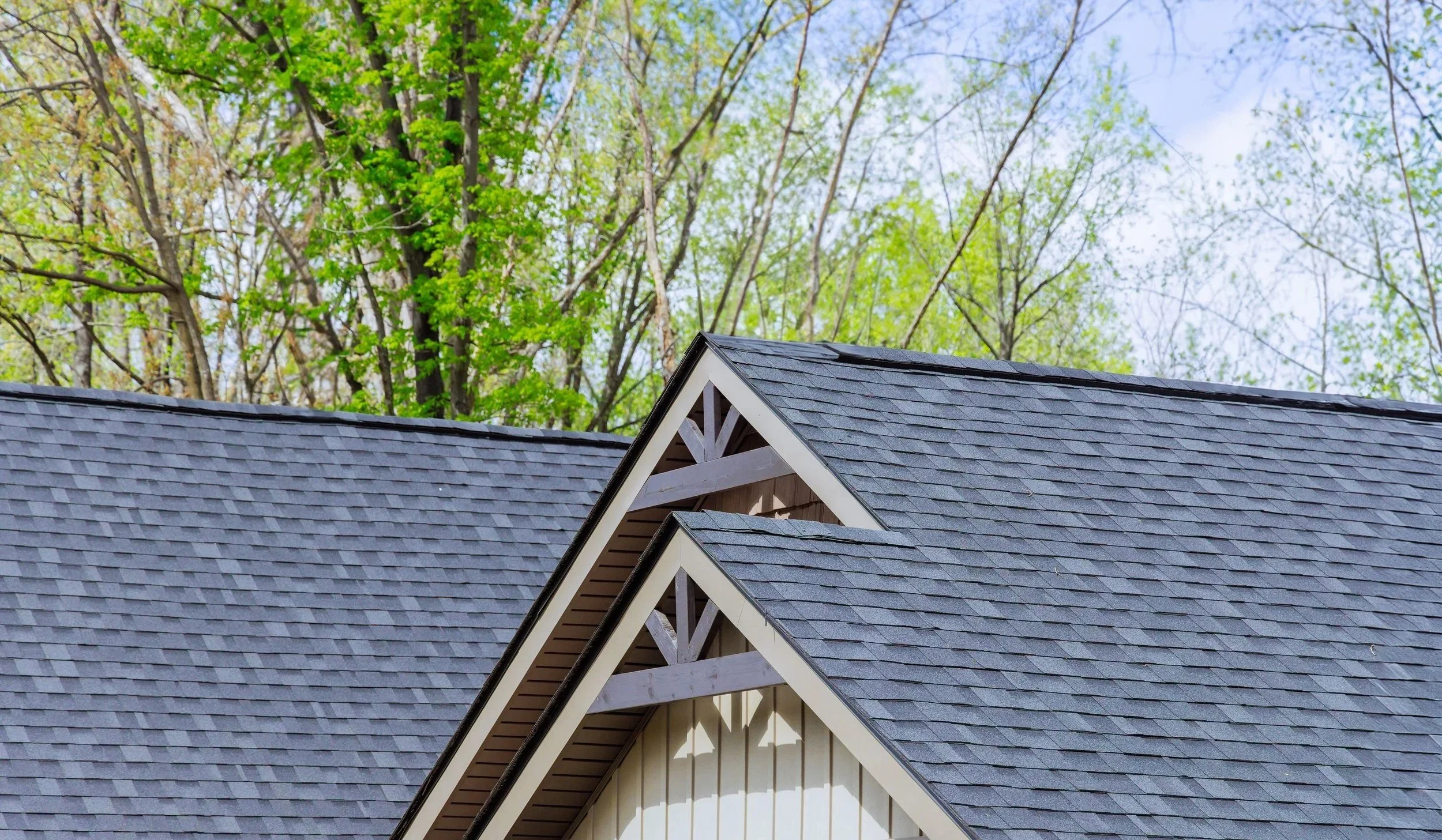 Close-up of a house roof with grey shingles and triangular gables, green trees in the background under a blue sky.