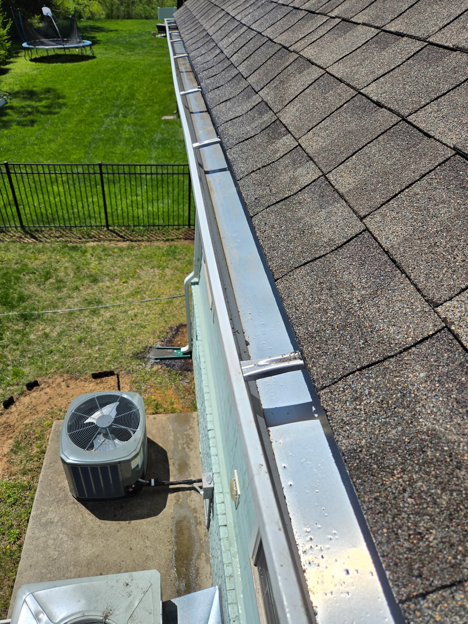 View from the roof showing asphalt shingles, gutter system, air conditioning unit, part of the house exterior, a fenced backyard with grass, a trampoline, and trees in the background.