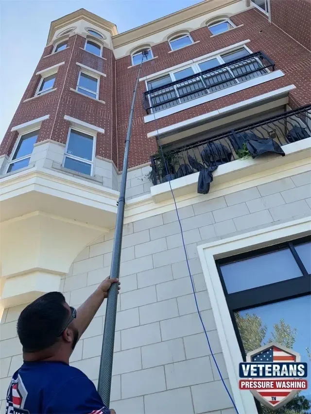 A man is pressure washing the exterior of a multi-story brick apartment building from the ground with a long pressure hose, cleaning the upper windows and walls.