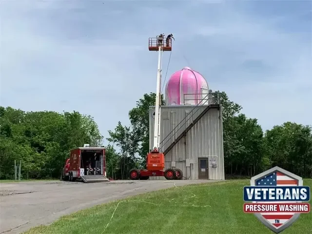 A pink observatory dome on a building with a cherry picker lift reaching the dome, surrounded by green trees and an open outdoor area. A vehicle is parked nearby with equipment, and a logo for Veterans Pressure Washing is visible in the corner.