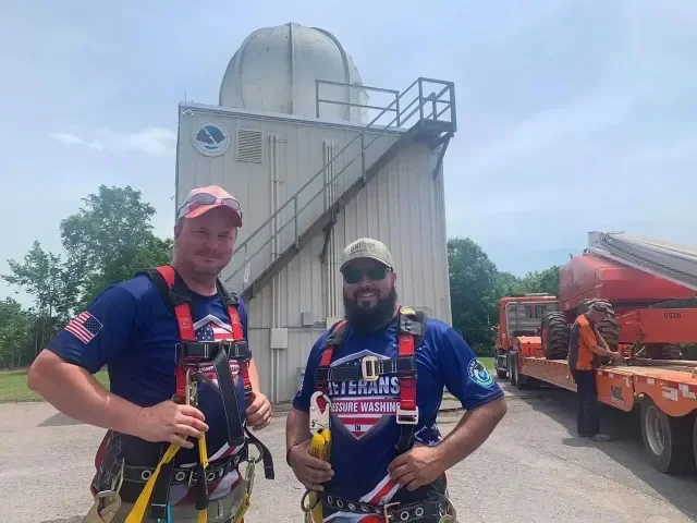 Two men in climbing gear standing outdoors near an observatory with a red and white truck in the background.