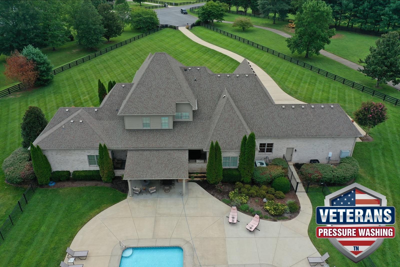 Aerial view of a house with a clean shingle roof, a swimming pool, a landscaped garden, and a driveway. There is a "Veterans Pressure Washing" logo in the bottom right corner.