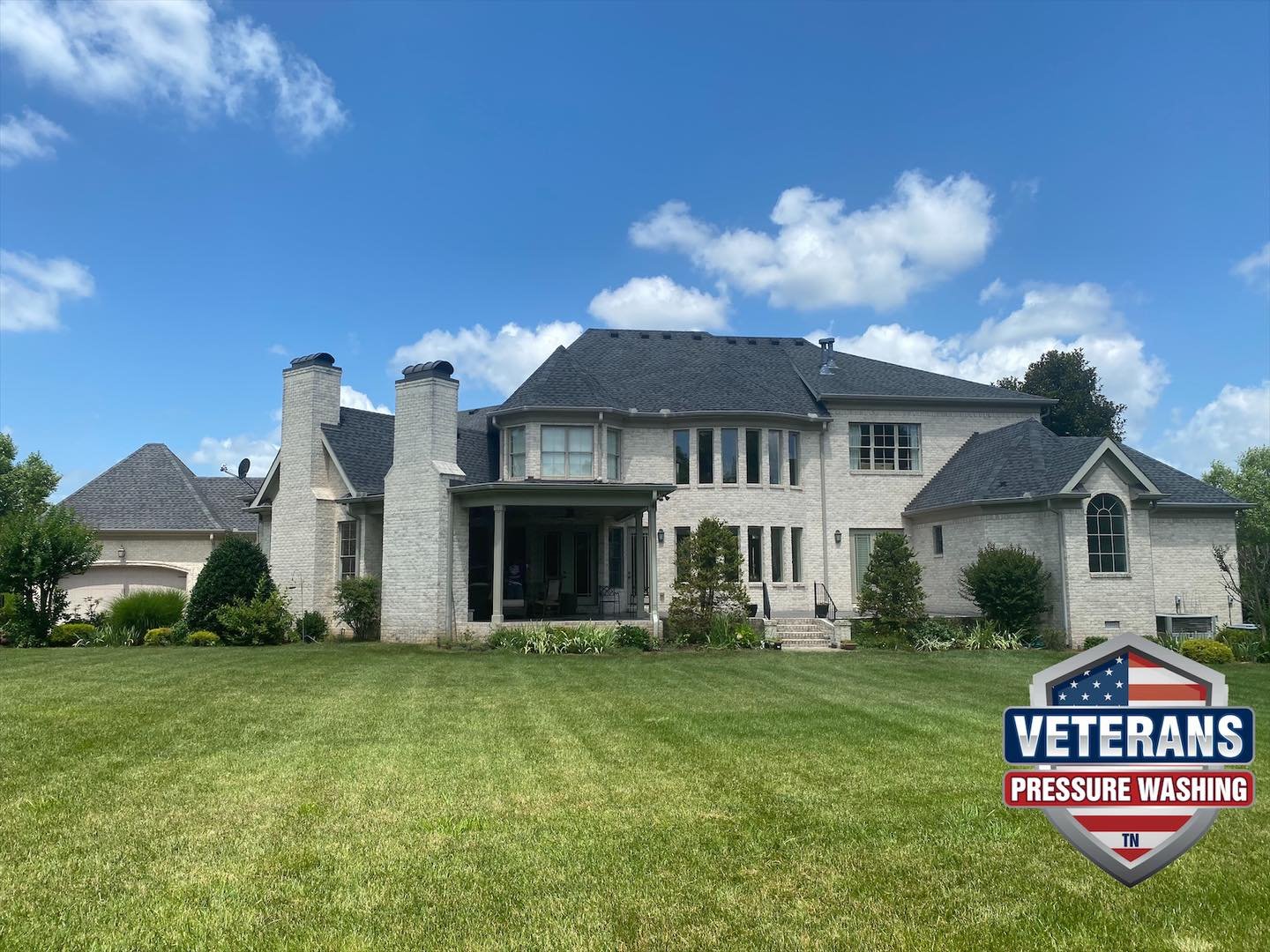 Large white brick house with multiple chimneys, a covered porch, and well-maintained lawn. A logo in the bottom right corner reads 'Veterans Pressure Washing' with an American flag design.