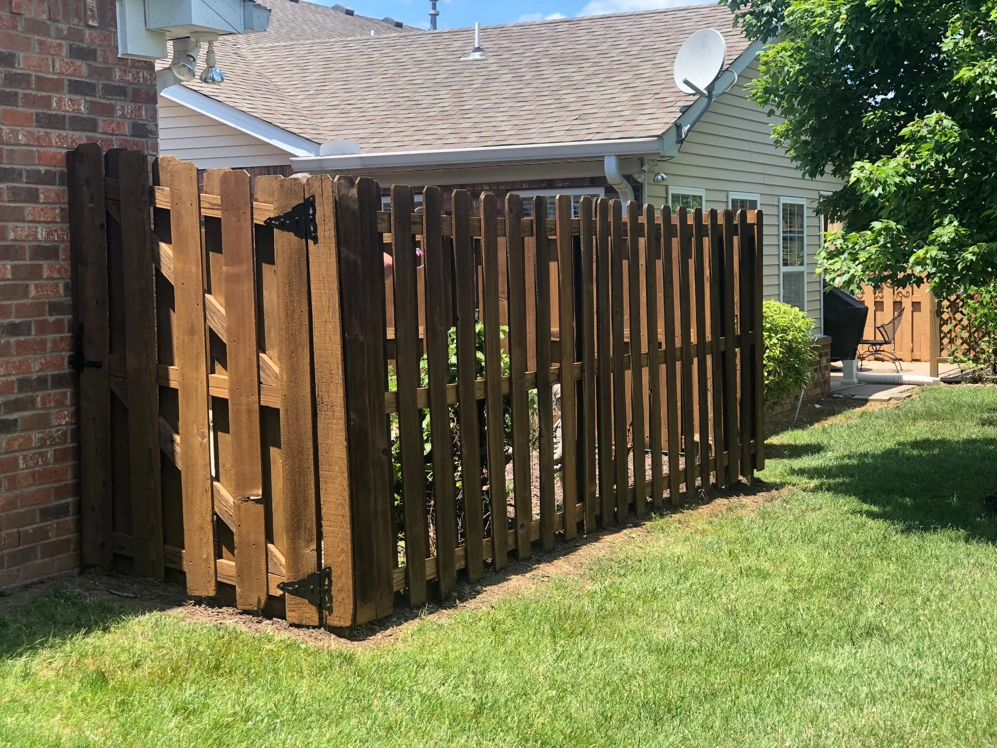 Wooden backyard fence with hinges near a brick house, green grass, and a tree with foliage. A house with beige siding, a satellite dish, and a patio with outdoor furniture are visible in the background.