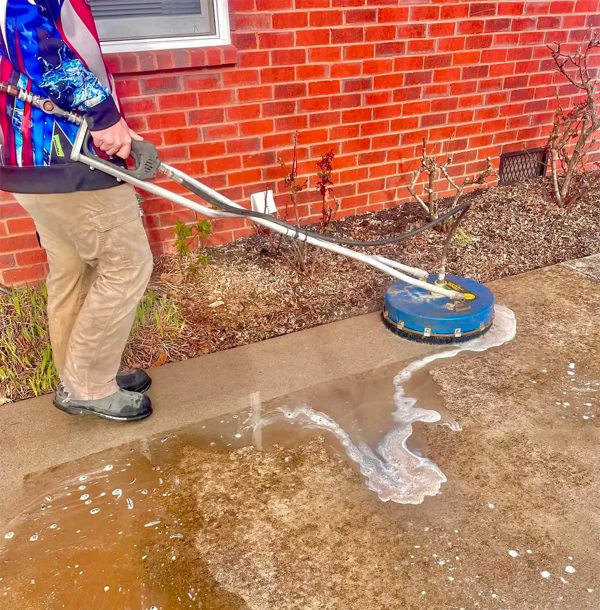 Person pressure washing a concrete porch in front of a brick house, with soap and water on the surface.