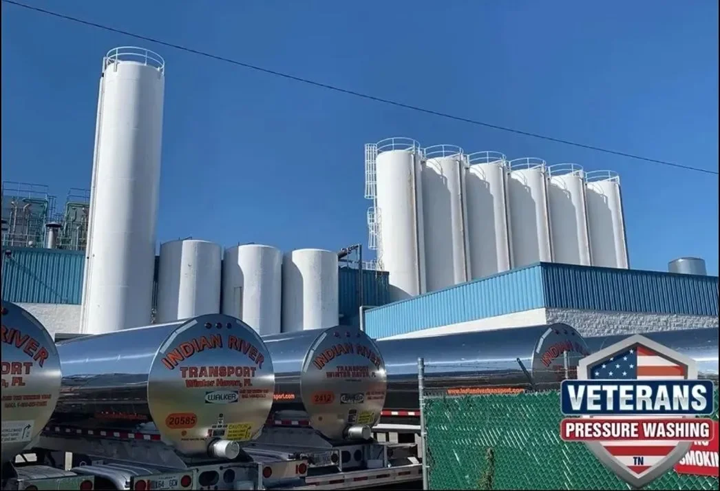 Industrial facility with large white storage tanks and silos, metallic pressure tanks labeled 'Indian River Transport' parked in front, blue building, green fence, and a hydration therapy logo reading 'Veterans Pressure Washing' in the lower right co