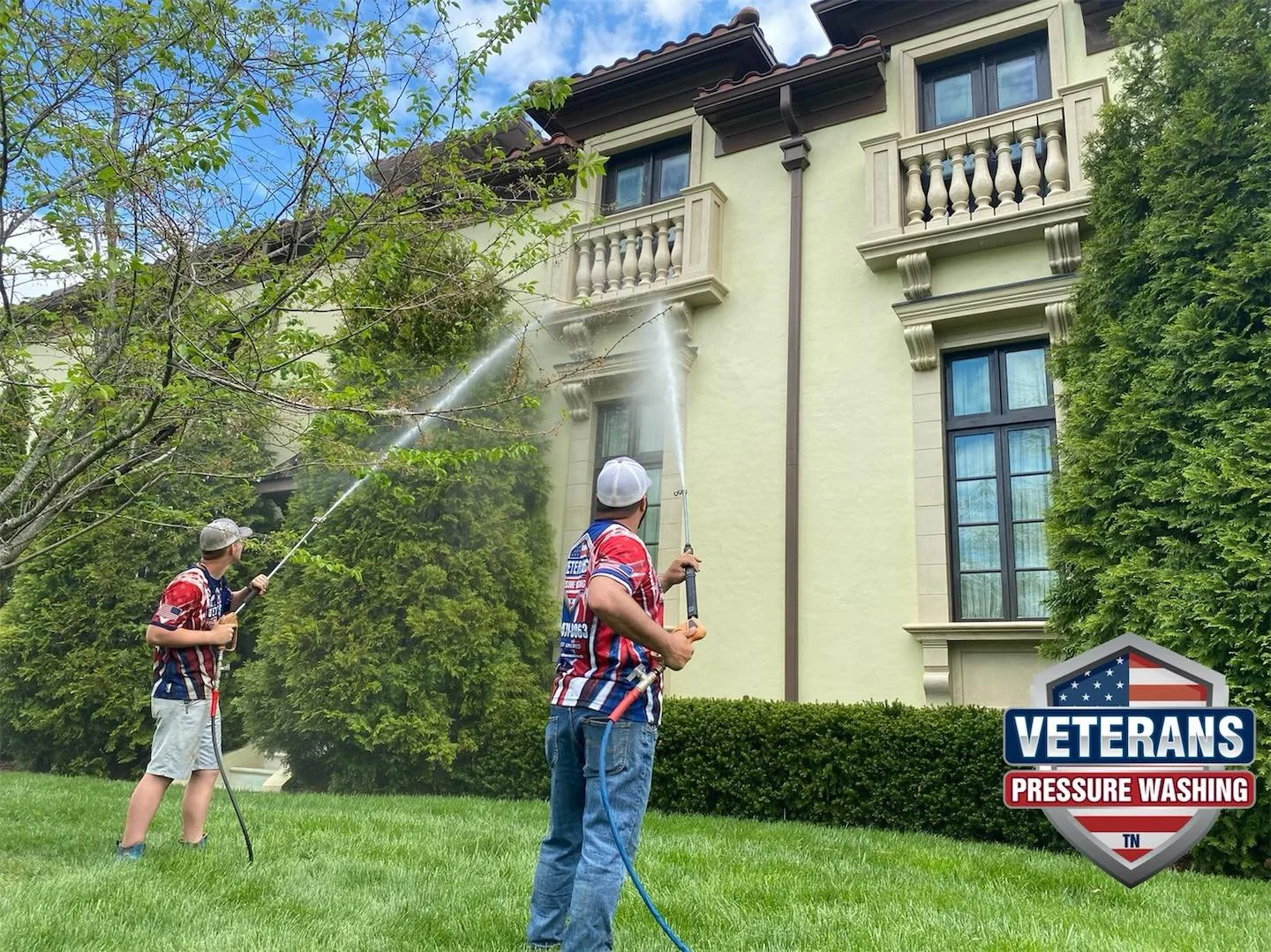Two men in patriotic shirts pressure washing the side of a large house with windows and balconies. The scene is outdoors on a green lawn with trees and bushes, under a blue sky with some clouds. There is a logo in the lower right corner that says ‘Veterans Pressure Washing, TN’.