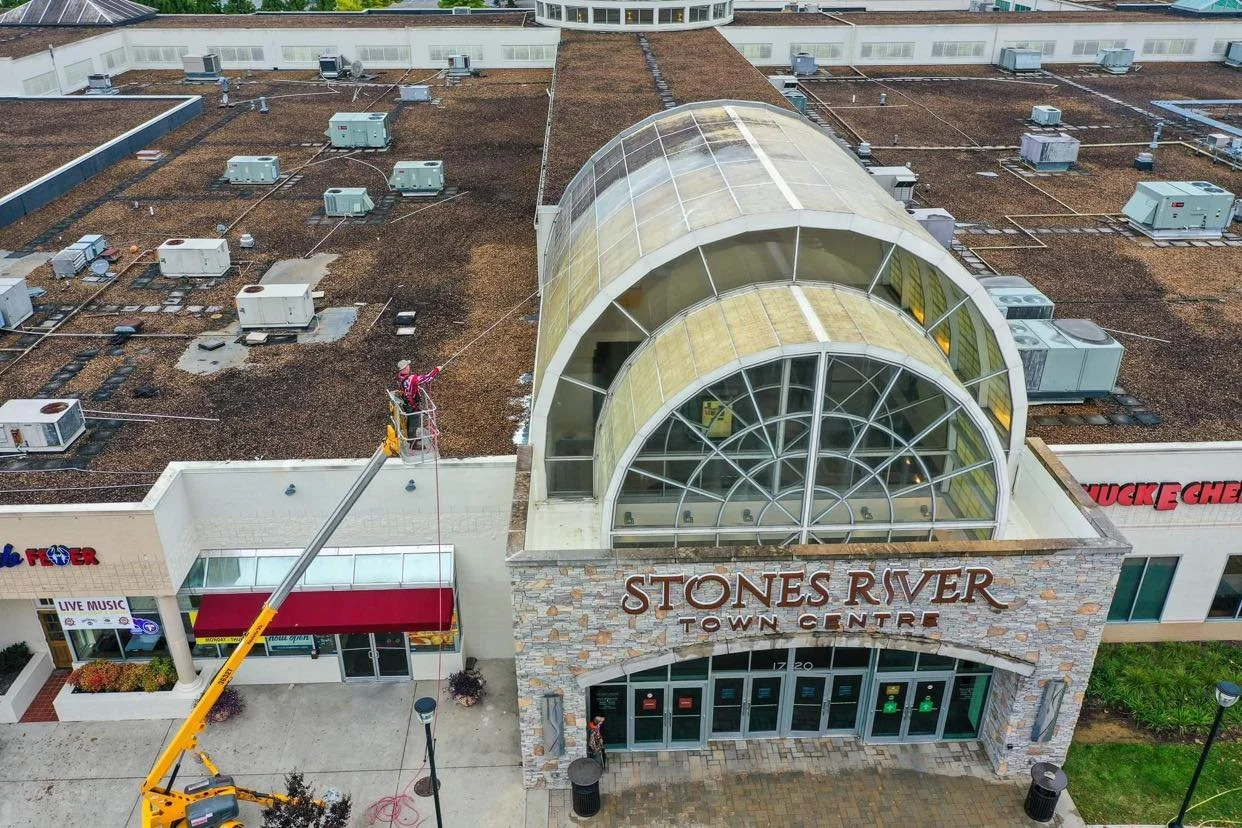 An aerial view of the Stone River Town Centre with a large glass-domed entrance and a worker on a crane cleaning the building's exterior.