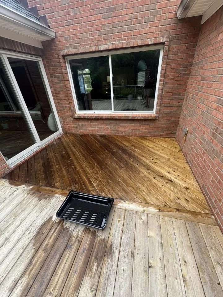 A small outdoor patio with brick walls and sliding glass doors, where part of the deck is being stained with wood stain. A black paint tray is placed on the lighter damaged wood section.