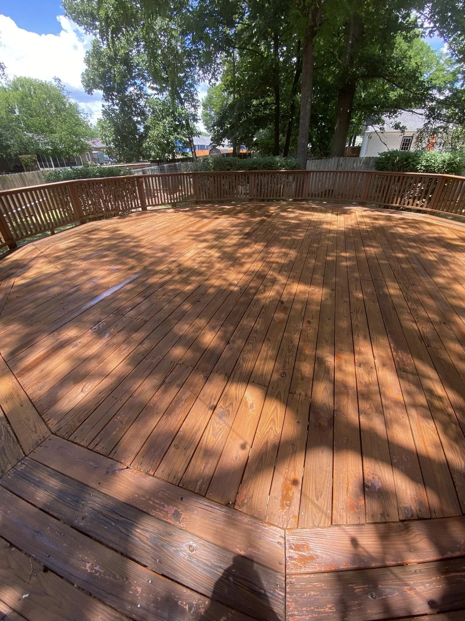 A wooden outdoor deck surrounded by trees and a wooden fence, with shadows cast by the branches and sunlight.