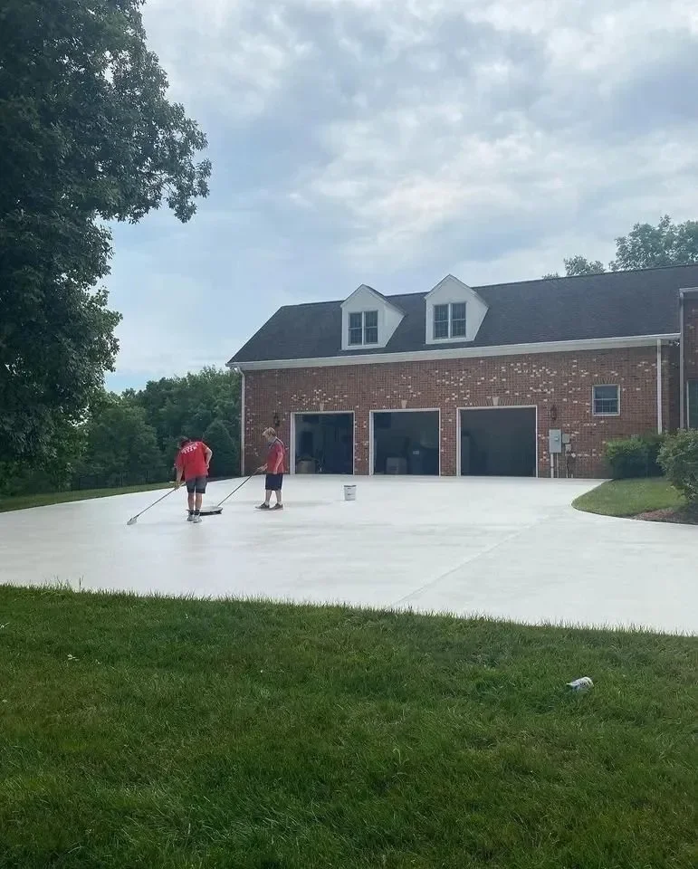 Two boys in red shirts are cleaning a newly poured concrete driveway in front of a brick house with a gray roof and three garage doors.