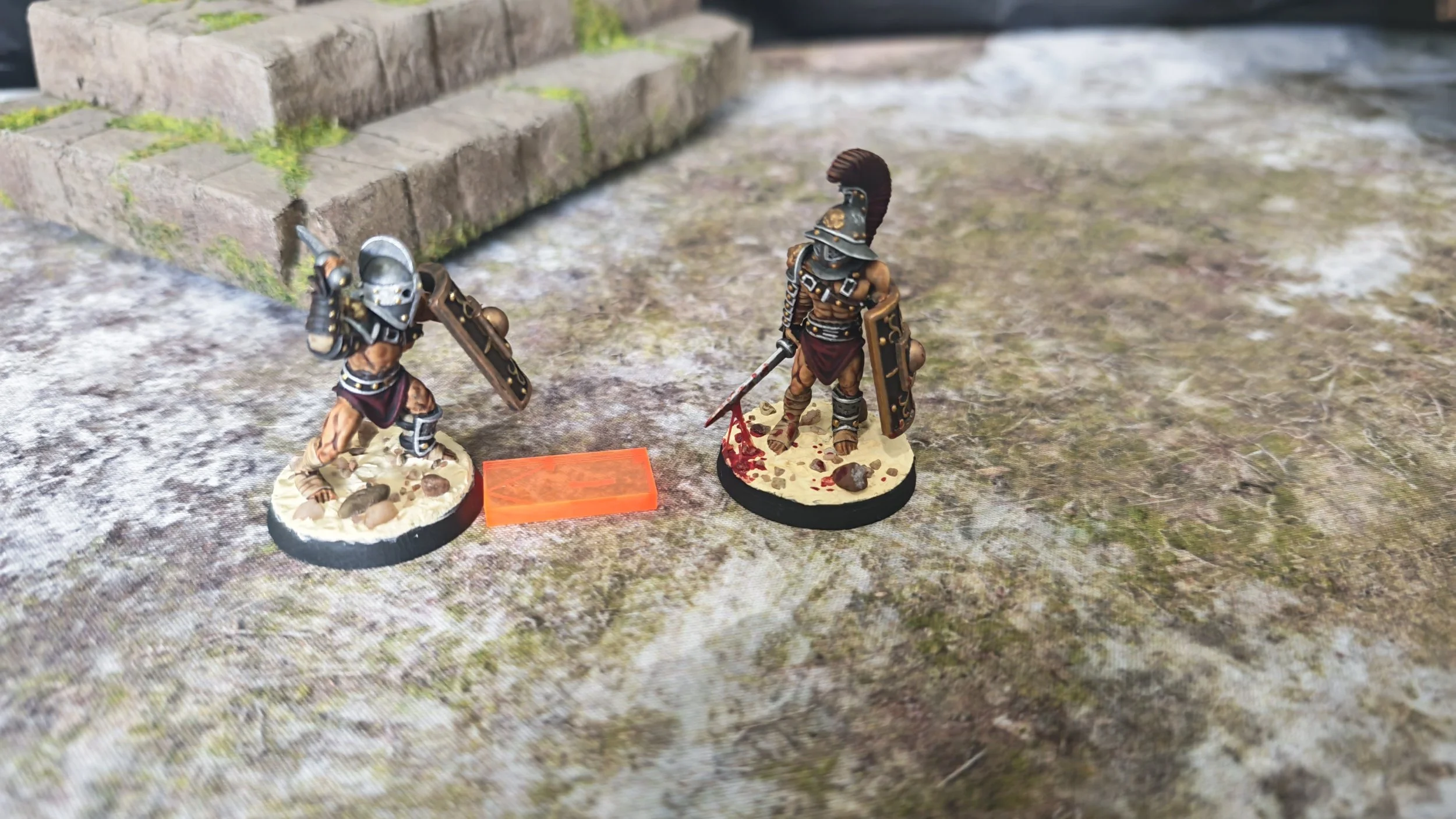 Two miniature figurines of ancient Greek or Roman warriors with shields and weapons, standing on textured bases, placed on a terrain-like surface near a stone step.