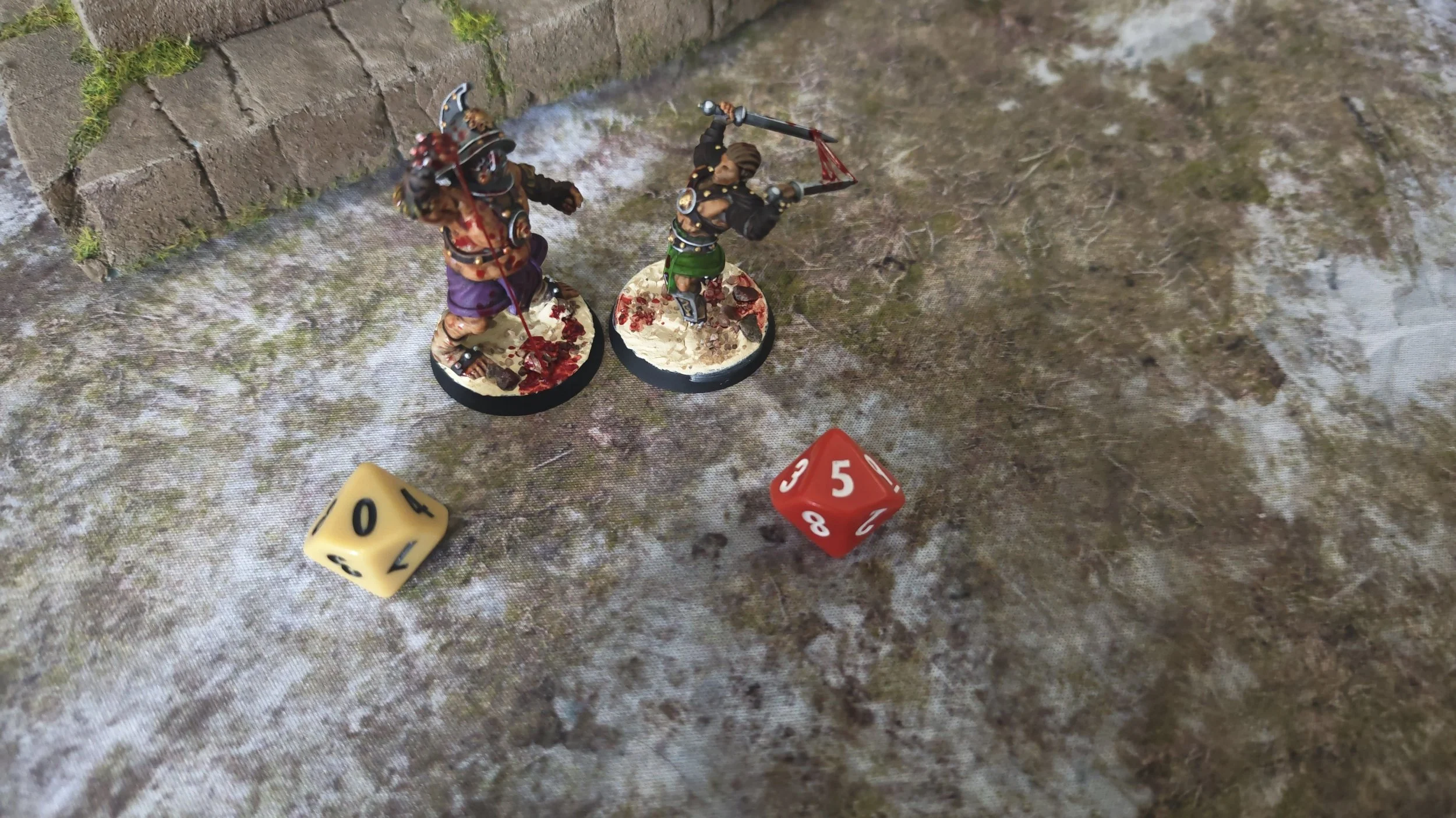 Two painted miniature warrior figurines engaged in battle on a textured surface, along with a yellow and red six-sided dice showing numbers 4 and 5.