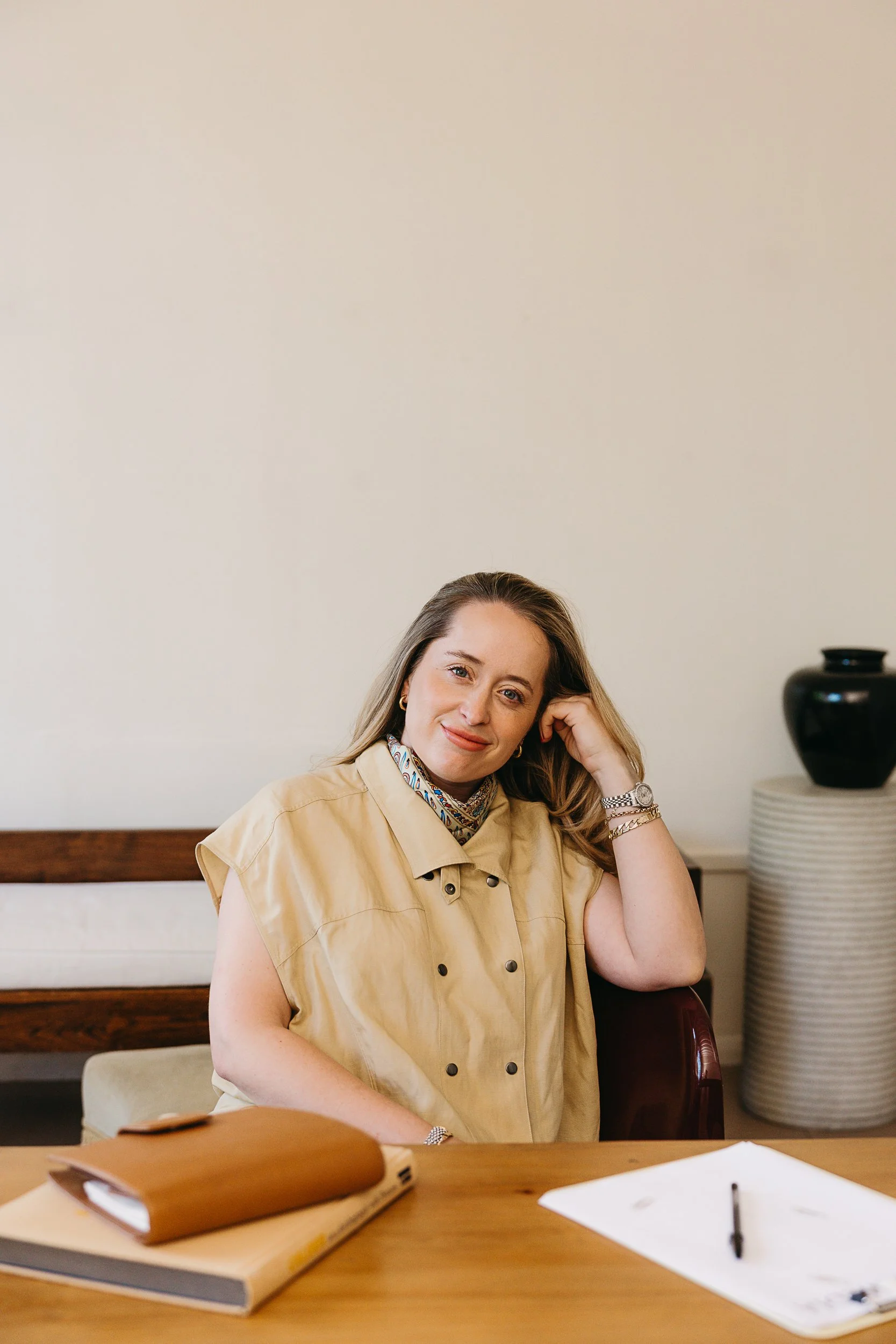 Woman with light brown hair, wearing a beige vest, sitting at a desk with notebooks, papers, and a pen. She is smiling and resting her head on her hand. In the background, there is a beige wall, a wooden ledge, and a black vase on a white side table.