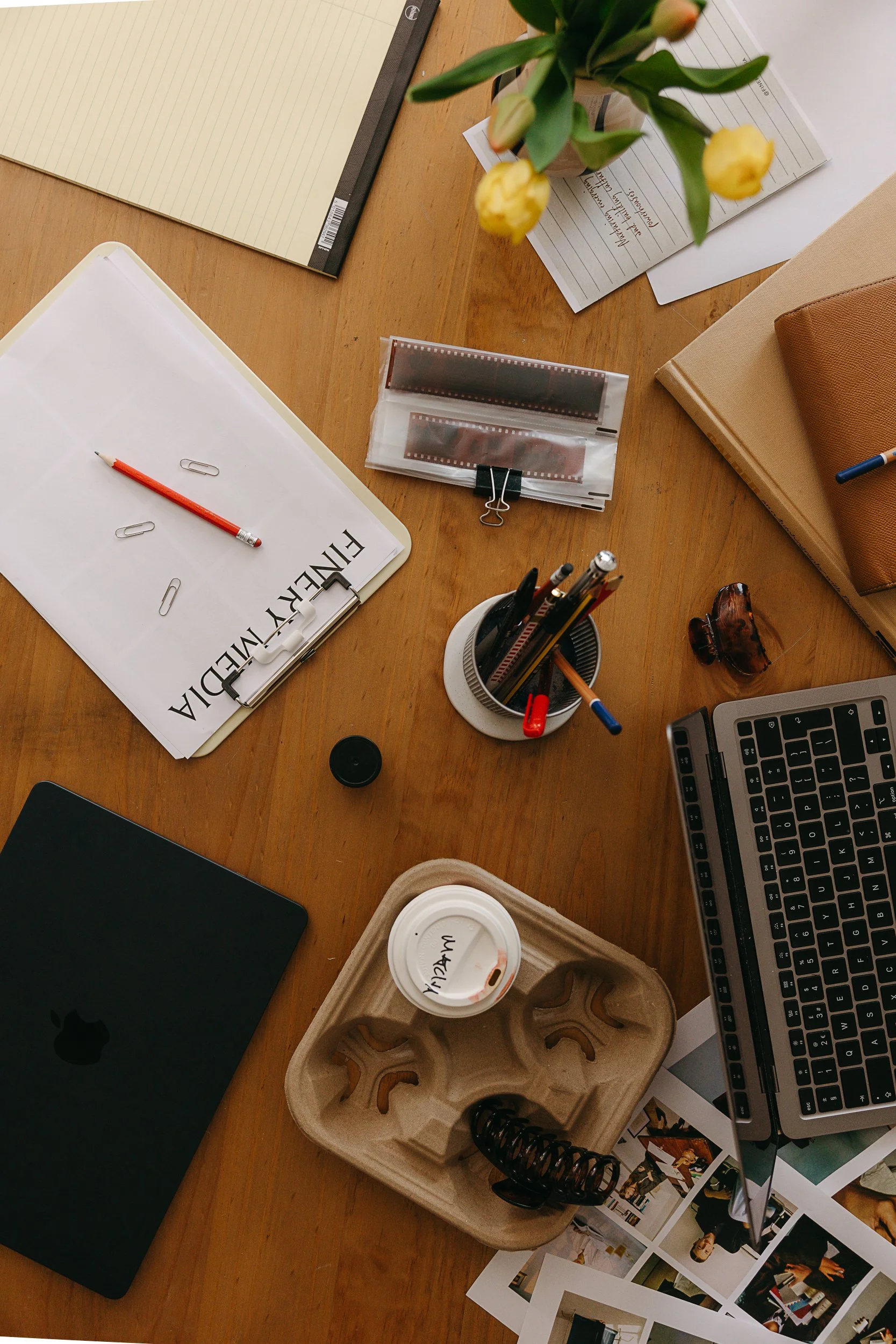 A cluttered wooden desk with various office supplies including notebooks, papers, a flower vase with yellow flowers, a black Apple computer, a coffee cup, clips, a film strip, and photographs.