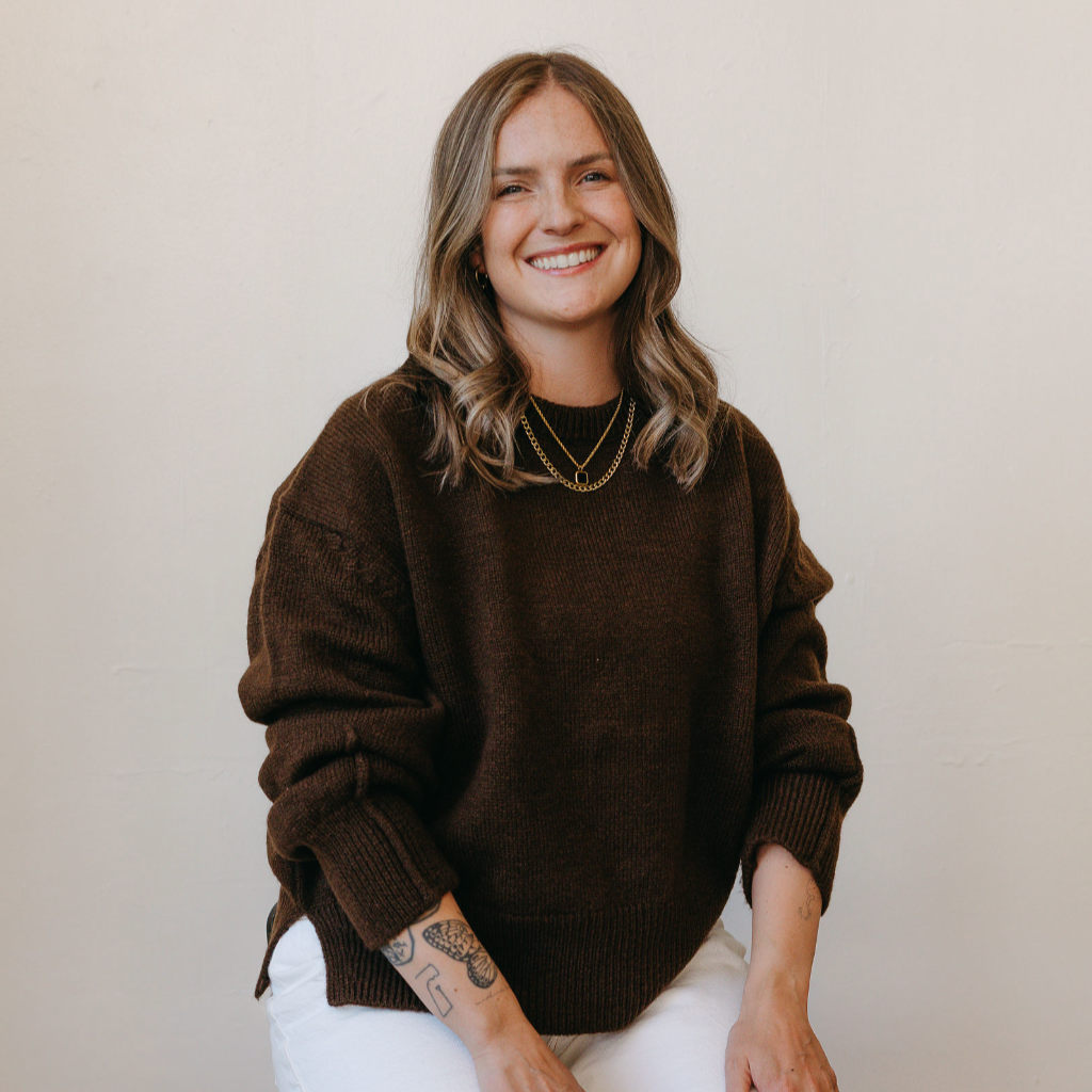 A young woman with shoulder-length wavy hair, smiling, wearing a dark brown sweater, white pants, and layered gold necklaces, sitting against a plain white wall.