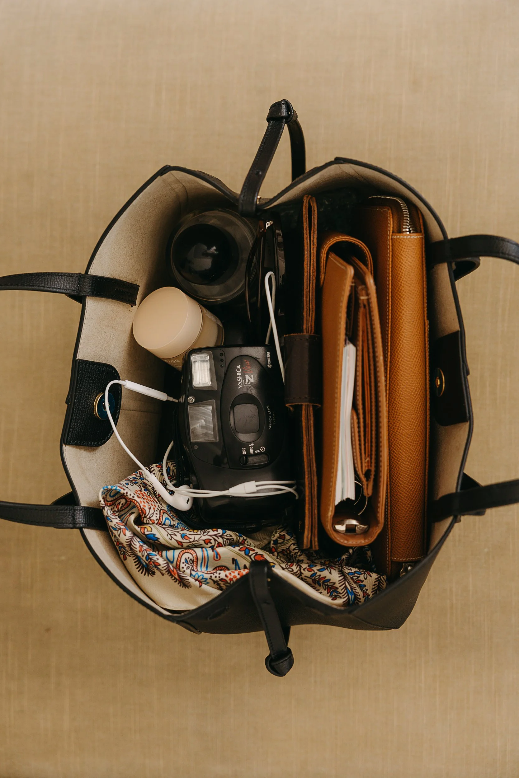 Top view of a black purse with various items inside, including a brown wallet, a small camera, a white candle, a colorful patterned scarf, a charger, and a brown notebook, against a light beige background.