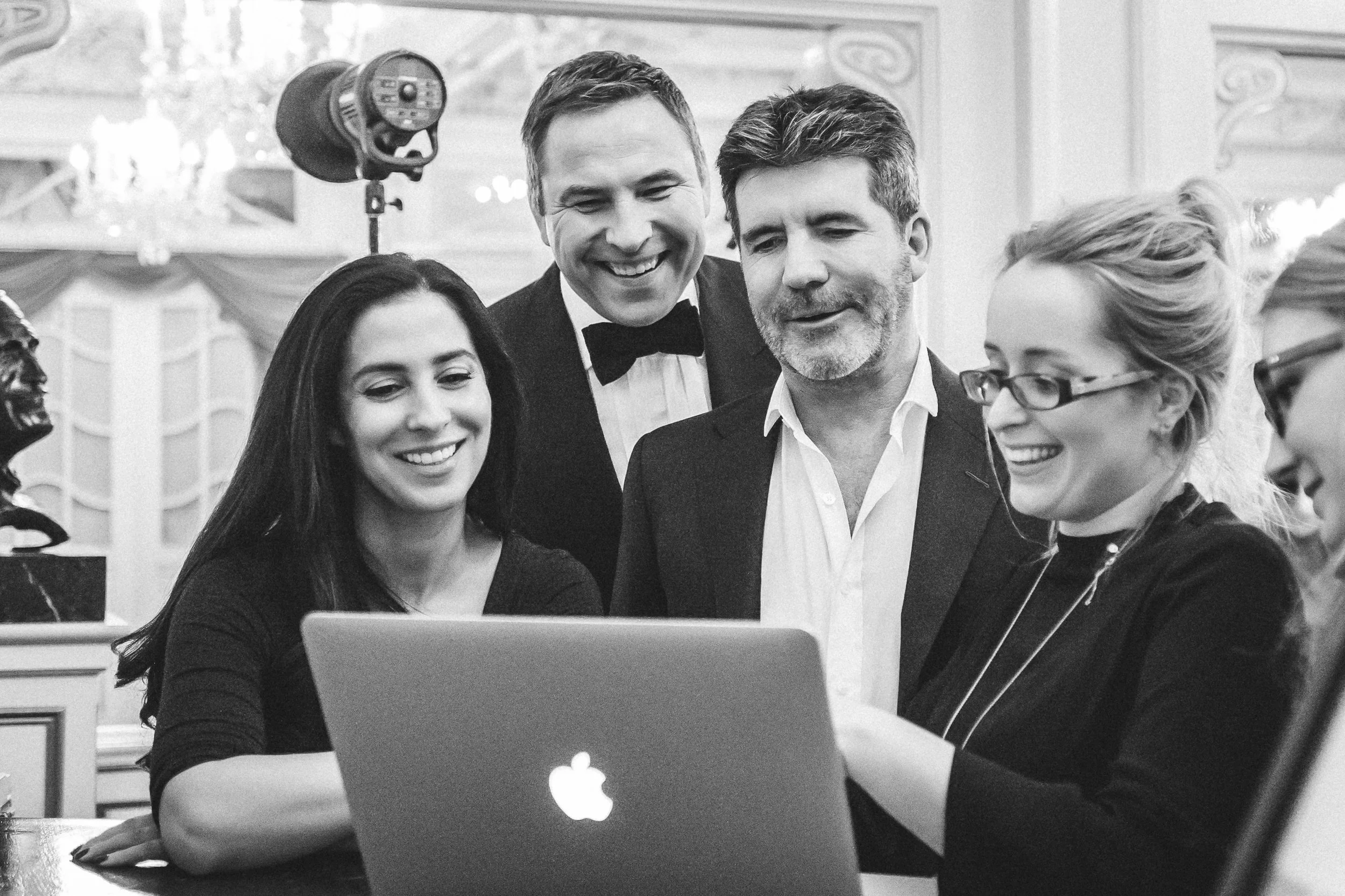 Group of four people gathered around a Dell laptop, looking at the screen, in an elegant room with ornate decor.
