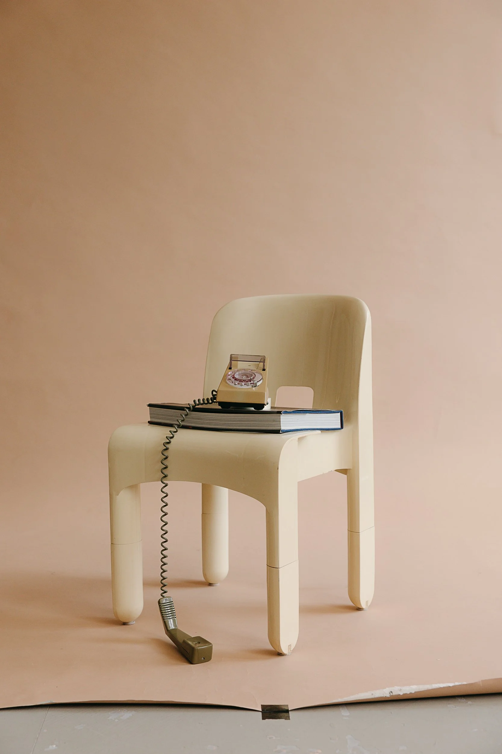 A vintage rotary telephone placed on top of a stack of books, sitting on a white chair against a plain beige wall in the Finery Media shoot.