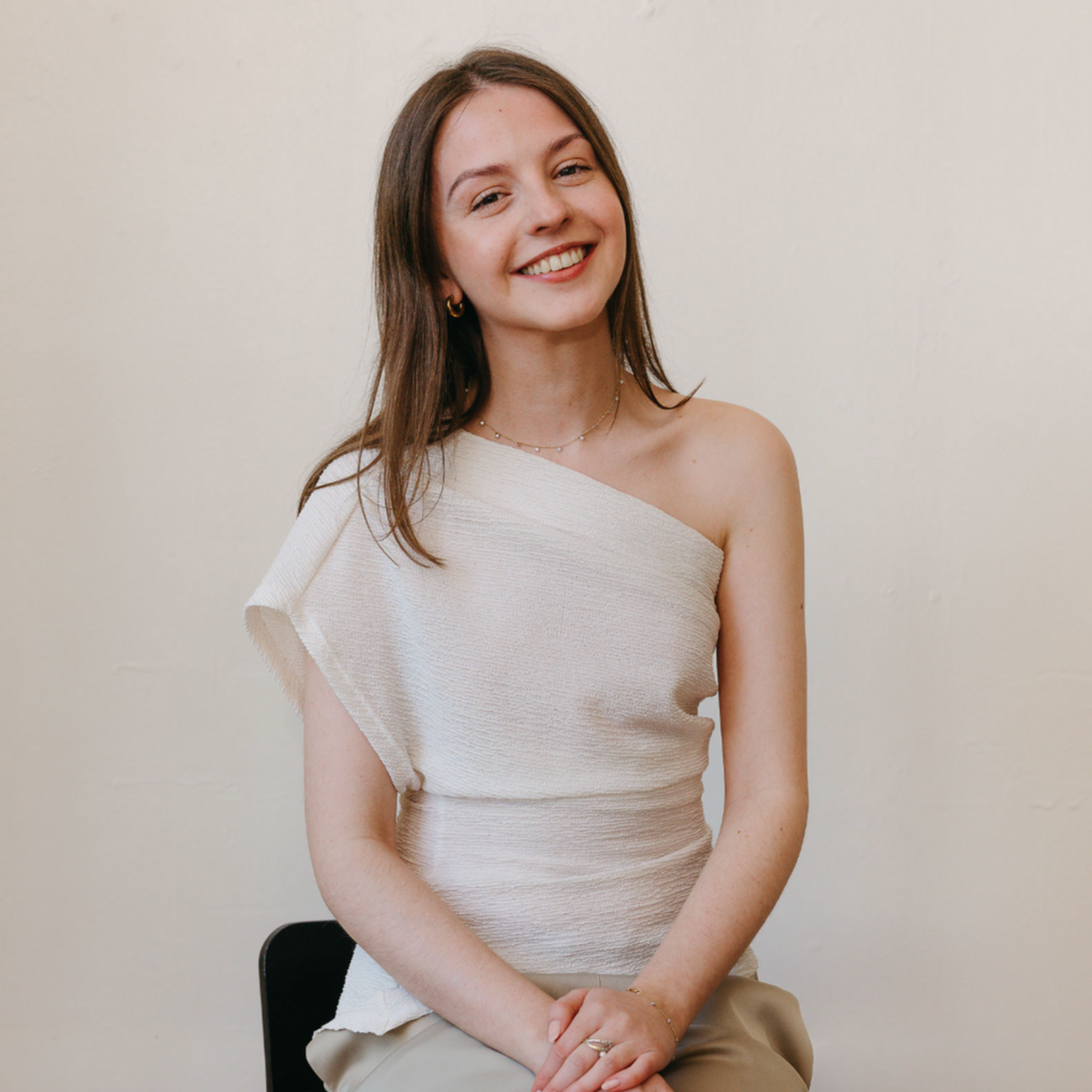 A young woman with long brown hair, smiling and looking at the camera, wearing an off-shoulder white top and beige pants, sitting on a black chair against a plain white wall.