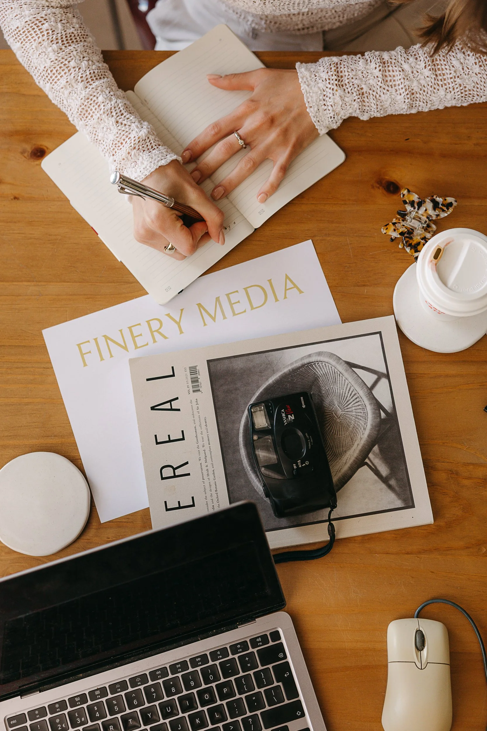 Finery Media imagery. Overhead view of a wooden desk with a laptop, a wireless mouse, a disposable coffee cup, magazines, a black case, and a person writing in a notebook. The person is wearing a white lace top and has a ring on their finger.