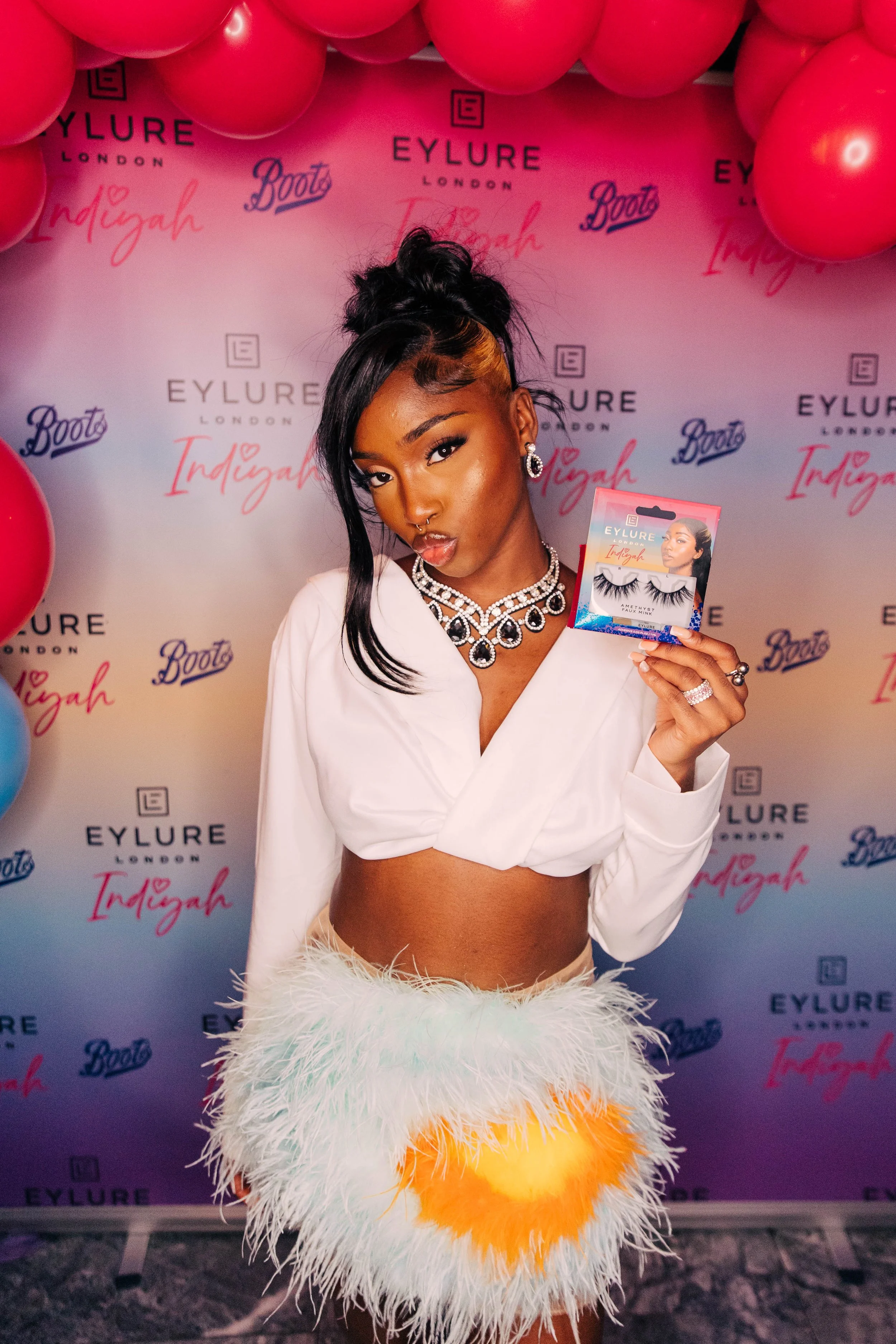 A woman with dark hair styled in a high bun, wearing a white blazer, a pearl necklace, and large earrings, holding a package of false eyelashes at an event with a pink and white backdrop with red balloons and logos for Eylure and Boots.