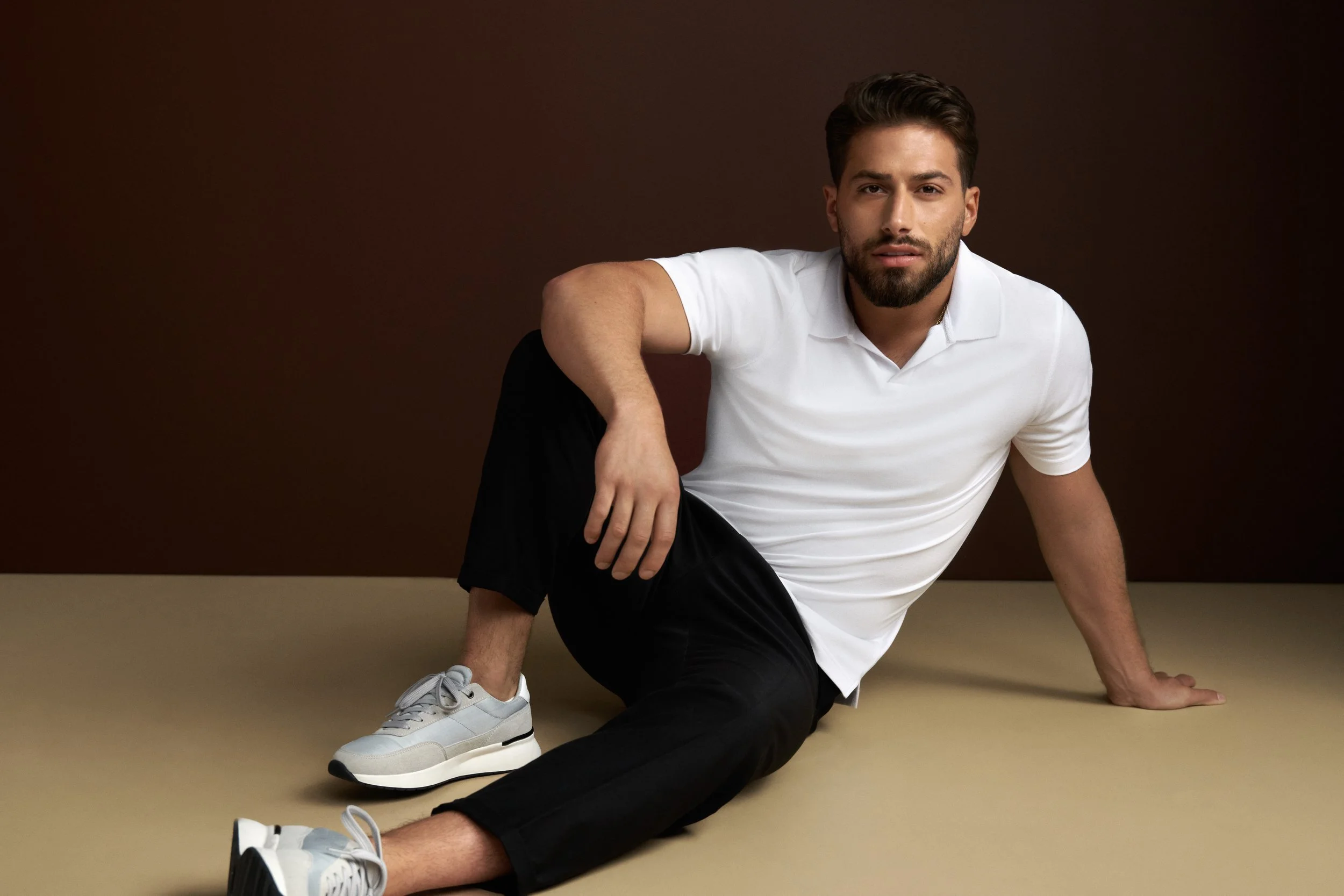 A man with short brown hair and a beard is sitting on the floor against a dark brown wall. He is wearing a white polo shirt, black pants, and white sneakers. He has one arm resting on his knee and the other arm supporting him as he leans slightly to the side.