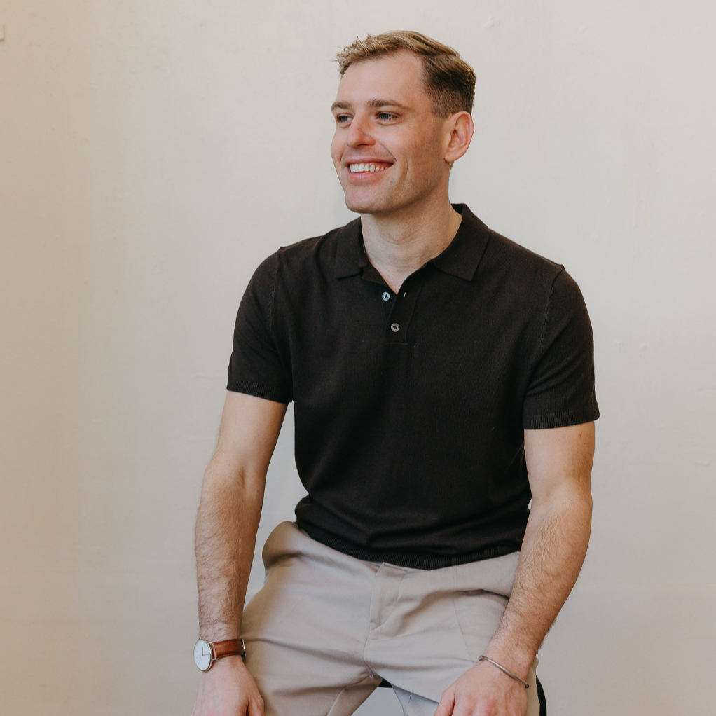 A young man sitting against a plain off-white wall, smiling and looking to his left. He is wearing a black polo shirt, beige pants, a brown watch, and a bracelet.
