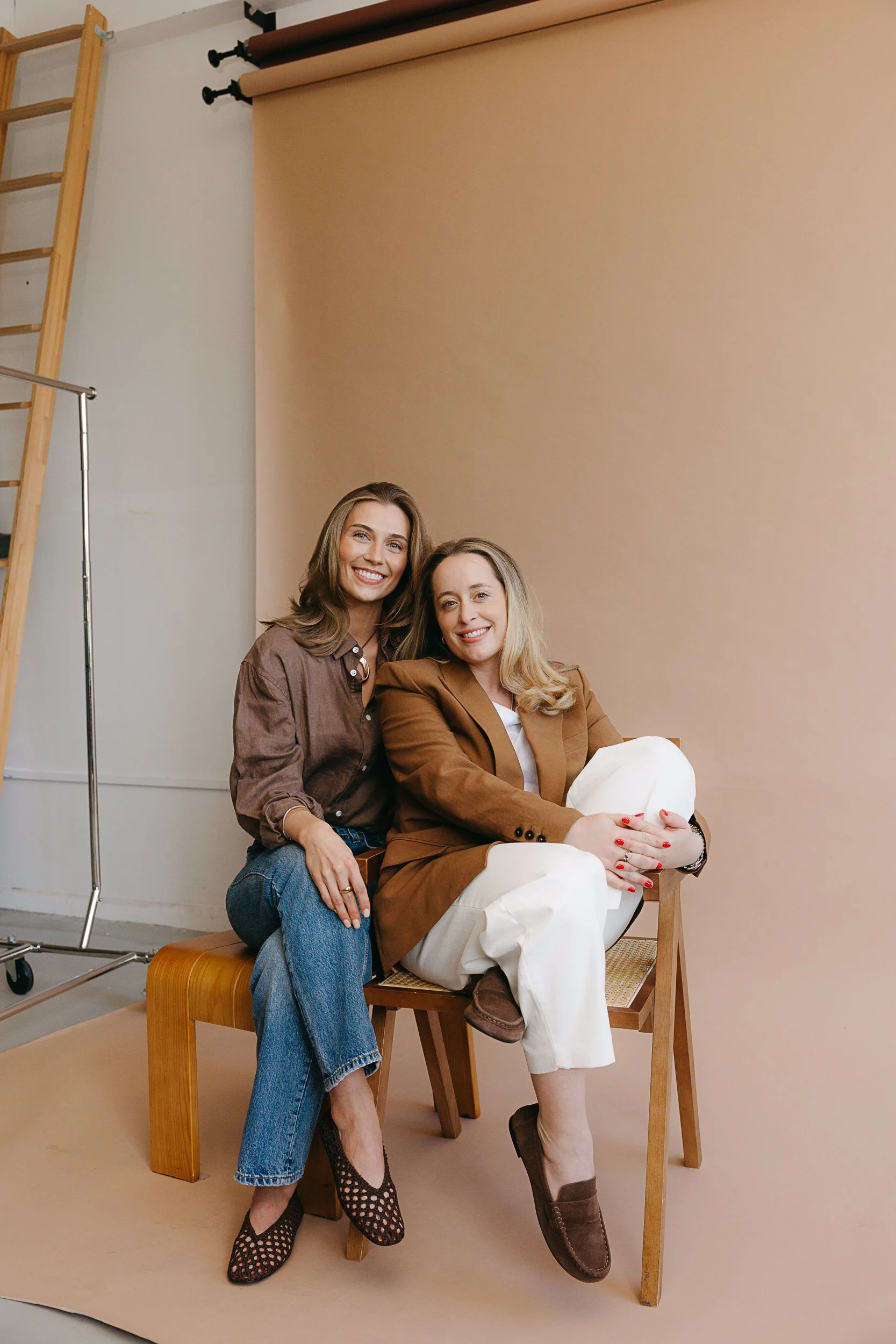 Two women sitting on a wooden chair in a photo studio, smiling, with a beige backdrop behind them. The woman on the left is wearing a brown shirt, blue jeans, and patterned shoes, with long brown hair. The woman on the right, seated, is wearing a brown blazer, white pants, and brown boots, and has blonde hair.