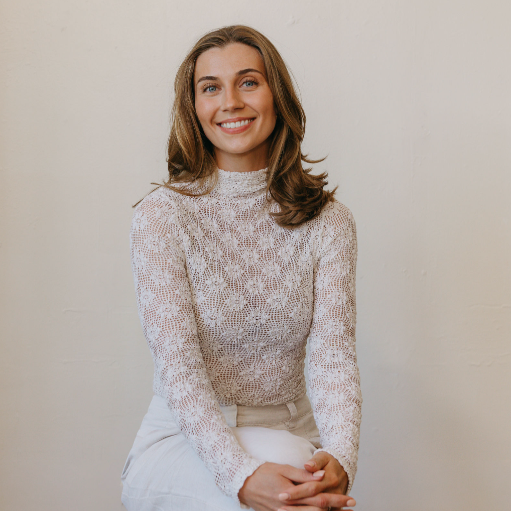 A young woman with brown hair and blue eyes sitting against a plain white wall, smiling, wearing a white lace long-sleeve top and white pants.